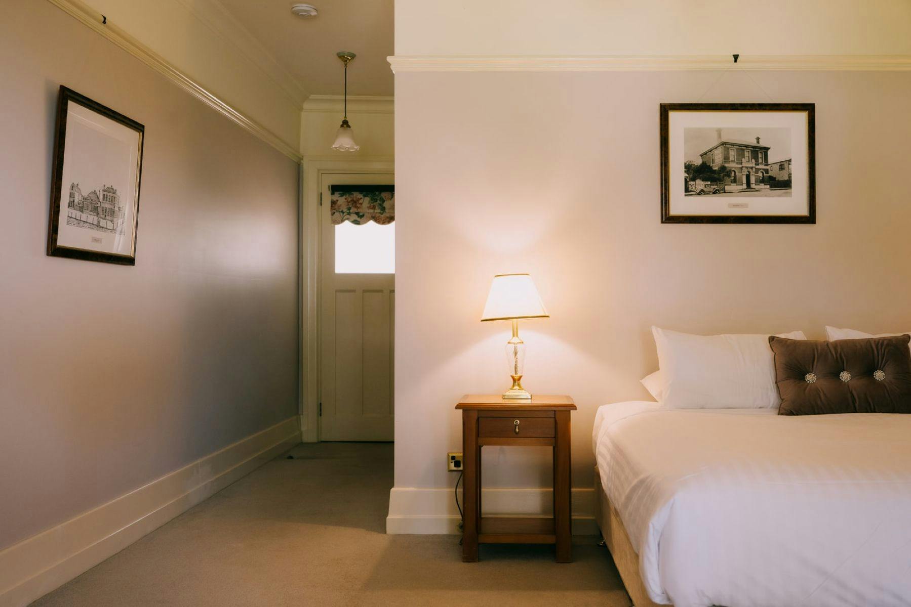 A cozy hotel room with a bed, nightstand, lamp, framed artwork, and a hallway leading to a door.