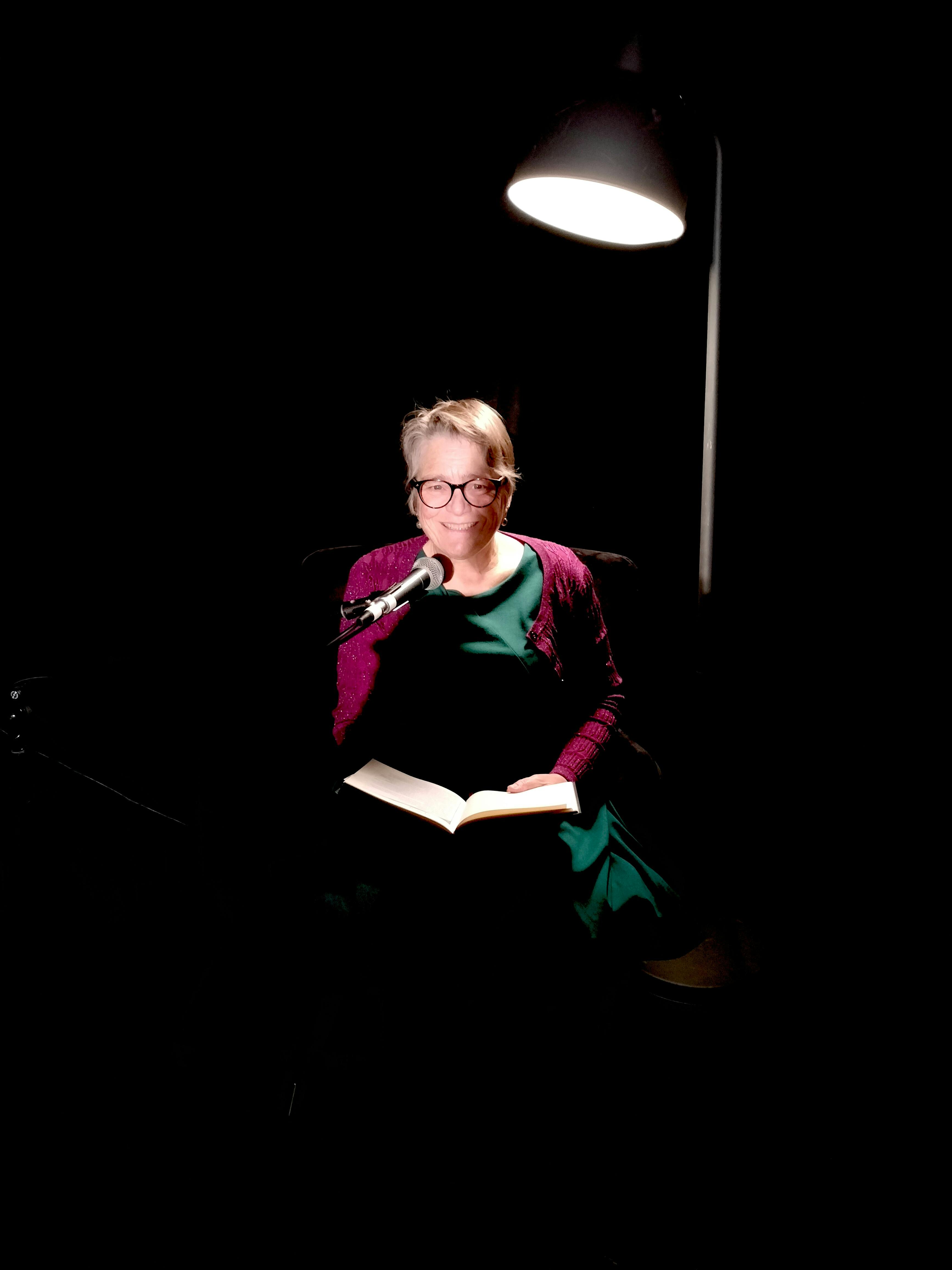 Woman in purple cardigan and green dress sat in chair and reading from an open book