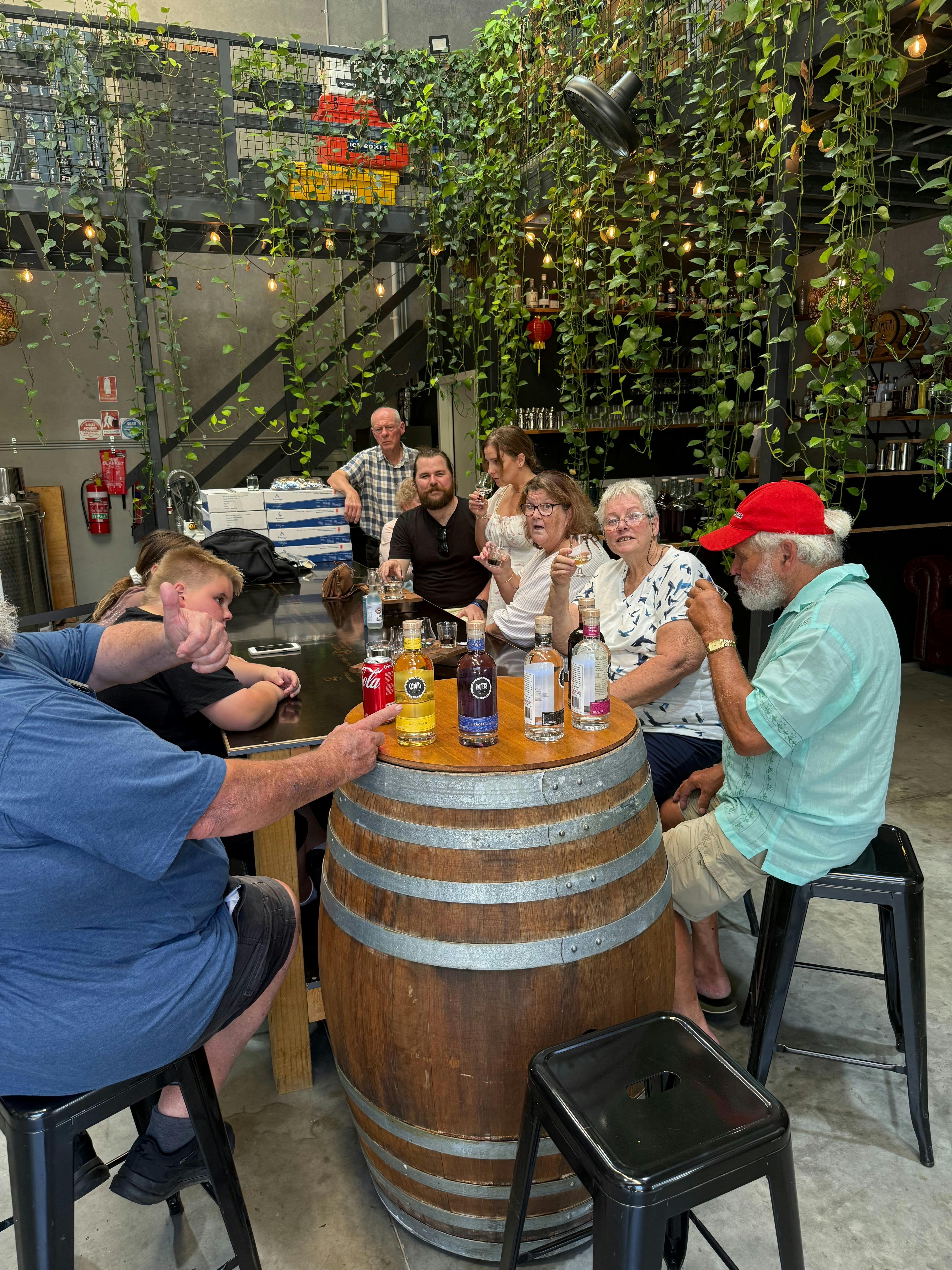 A group of guests gathered around a wooden barrel table displaying spirit bottles and tasting glasse