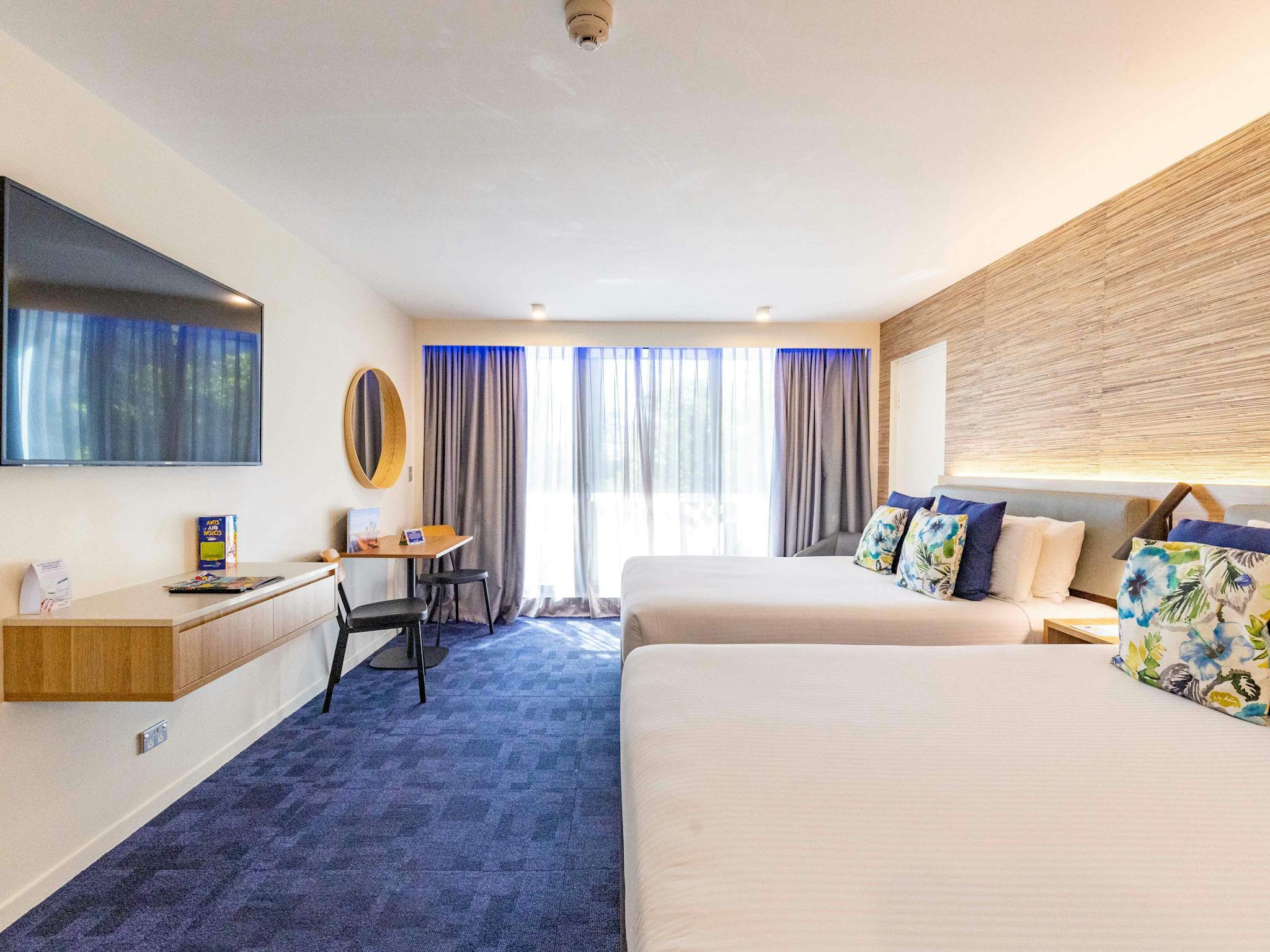 A room with dark carpet and two Queen beds with TV, shelf and glass doors and a sheer curtain A room with dark carpet and two Queen beds with TV, shelf and glass doors and a sheer curtain