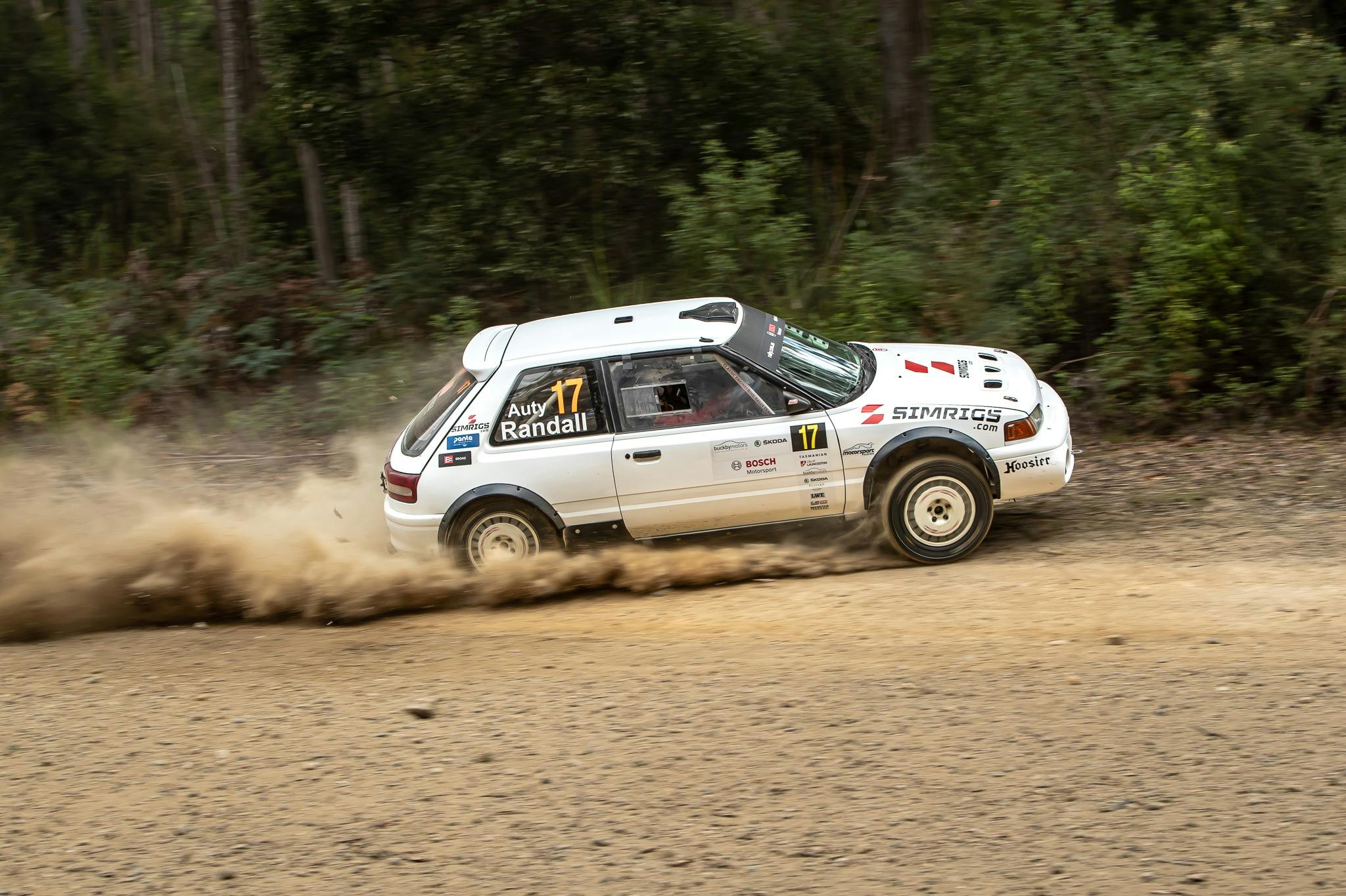 A white rally car kicking up dust as it turns a corner on a gravel road