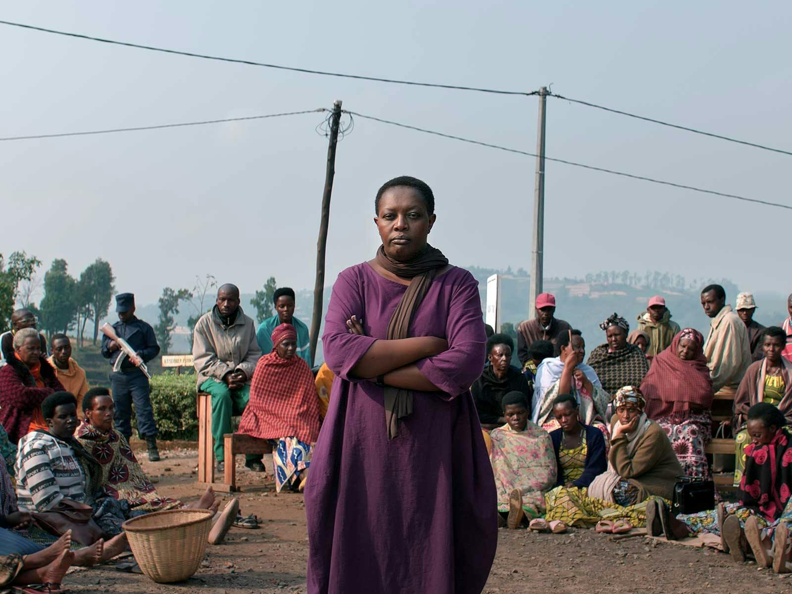 A woman in a purple dress stands in front of a crowd with a serious expression