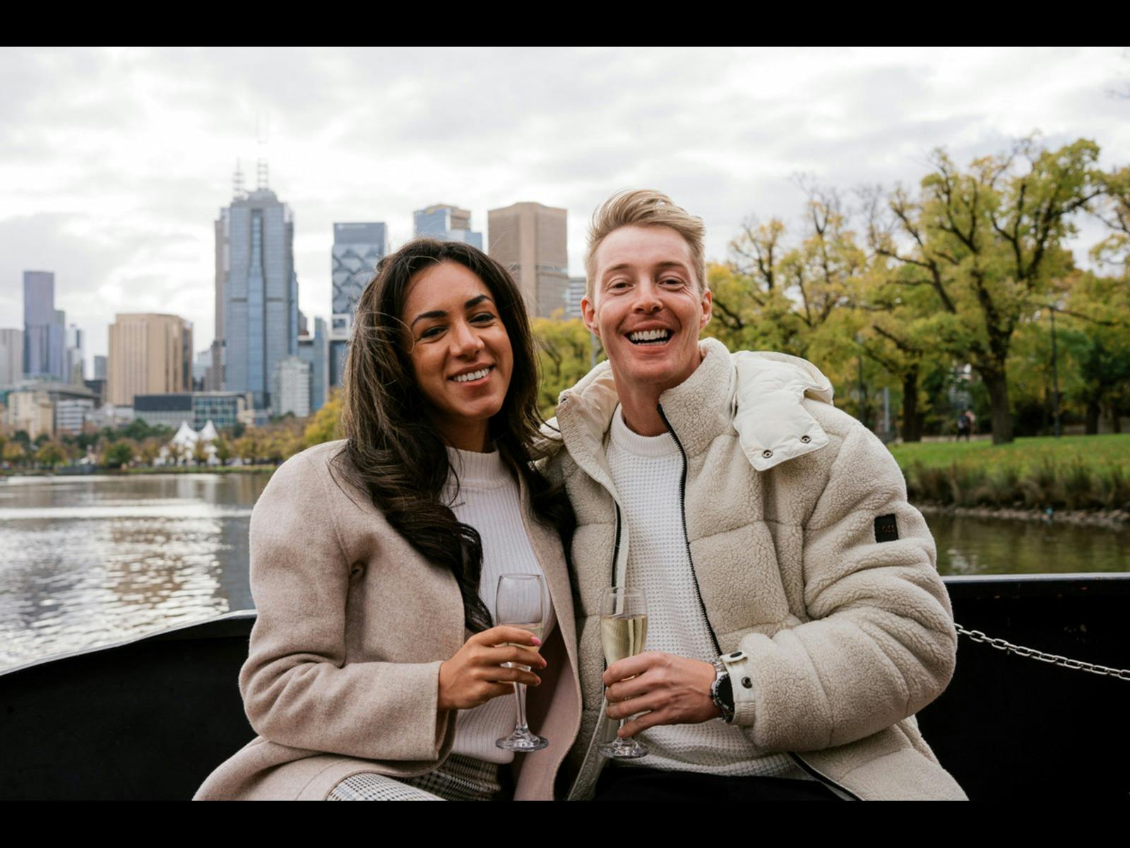 2 guests holding sparkling wine, sitting on the outside deck of cruise with city views behind them.