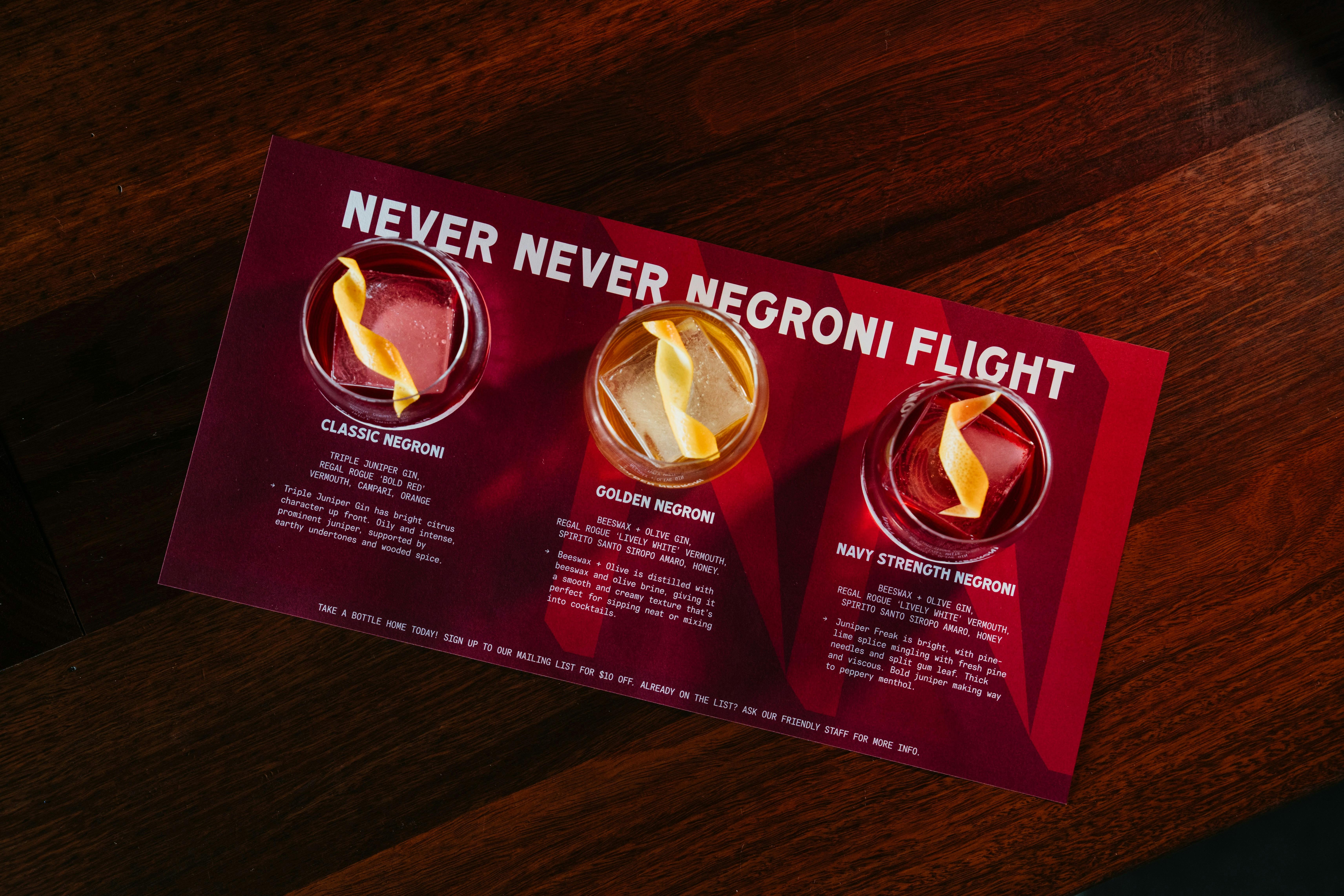 Three miniature negronis are served on a maroon tasting mat, with white text.
