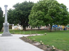A tall imposing photo of the war memorial in the Penola Park with playground in the background.