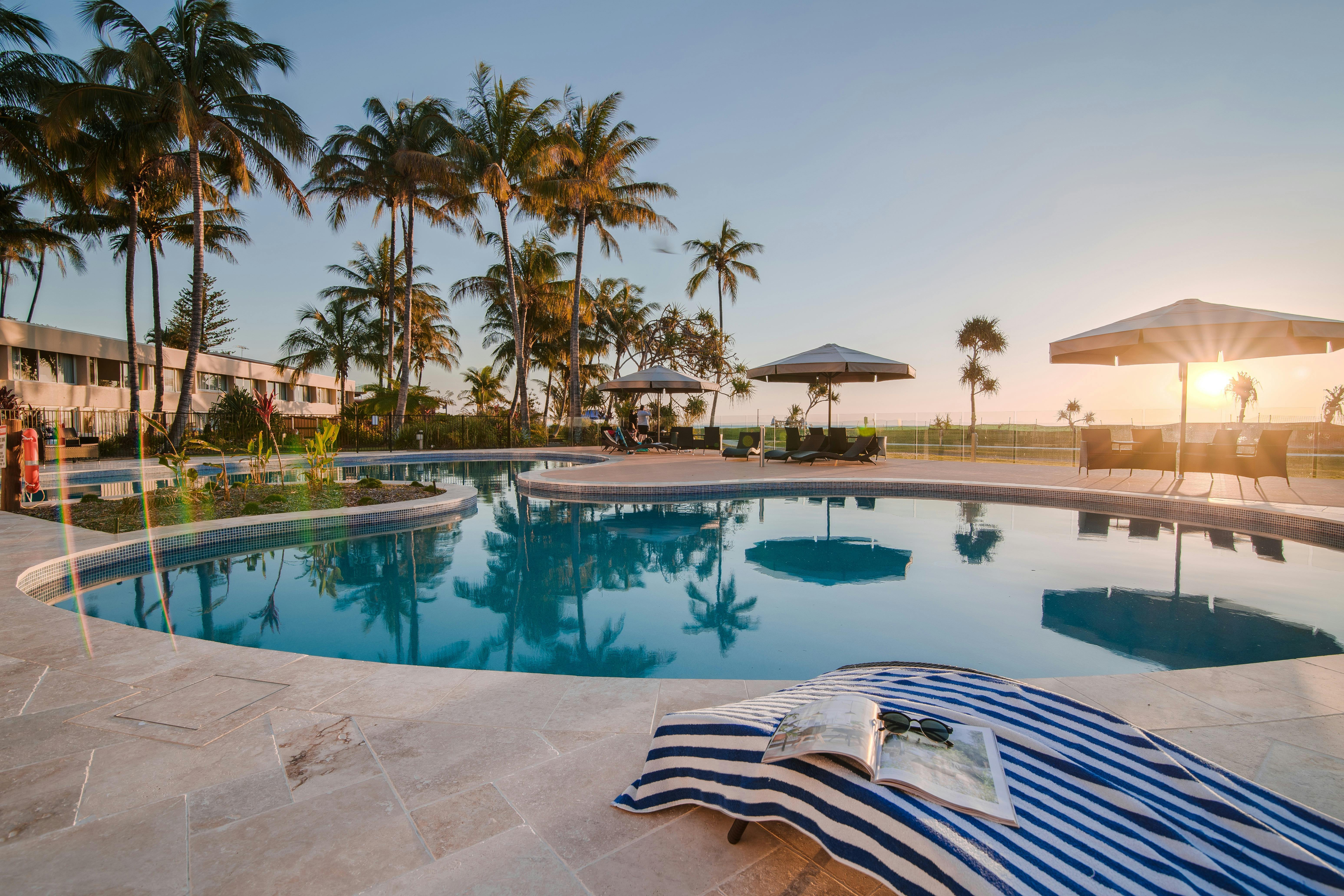 Pool at sunset with towel and book on lounge chair and palm trees in background