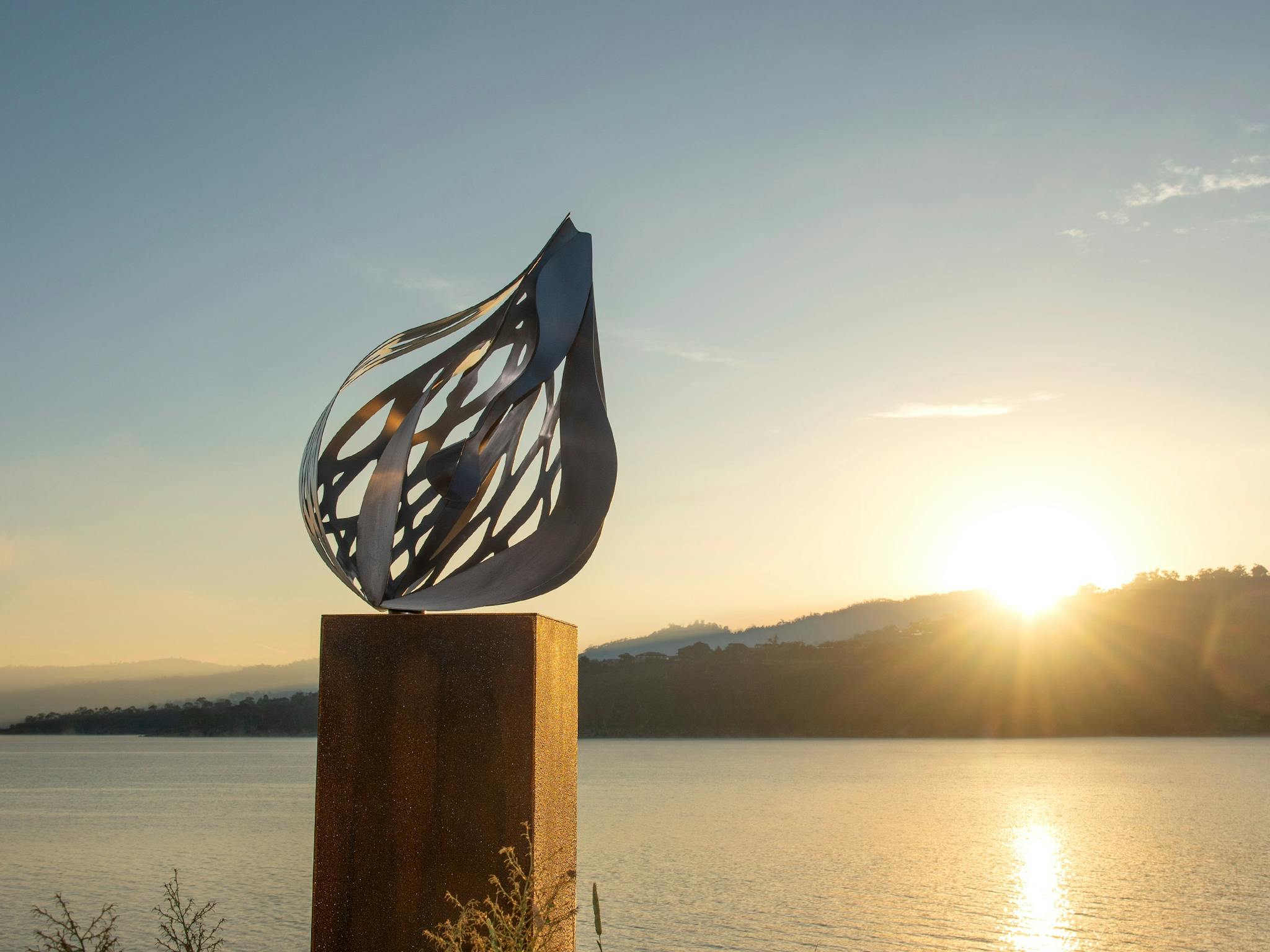 Metal sculpture of an intricate pod on a plinth, set against a still lake at sunrise.