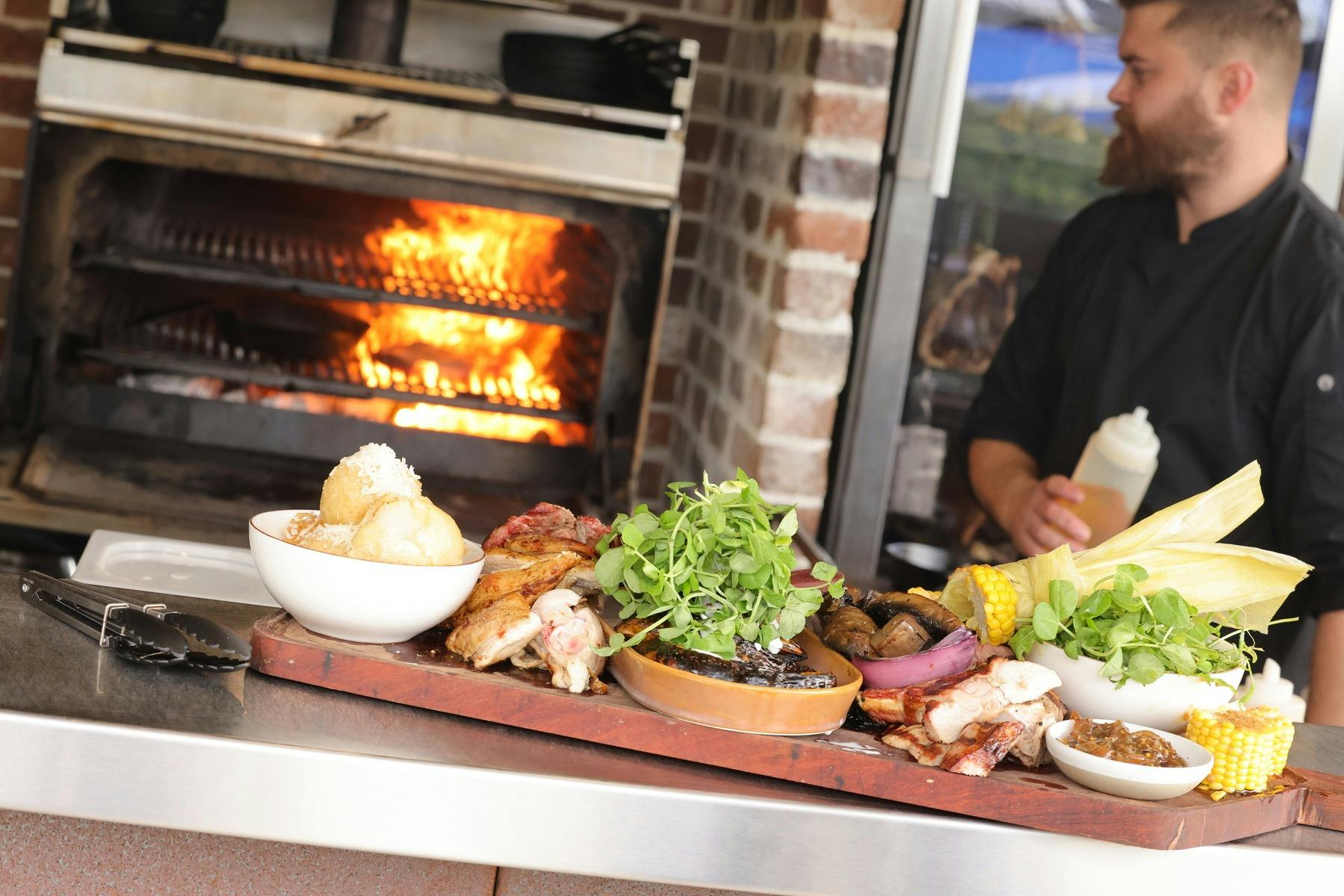 火焰烹調的食物，背景是廚師在 Mibrasa 明火烤箱旁