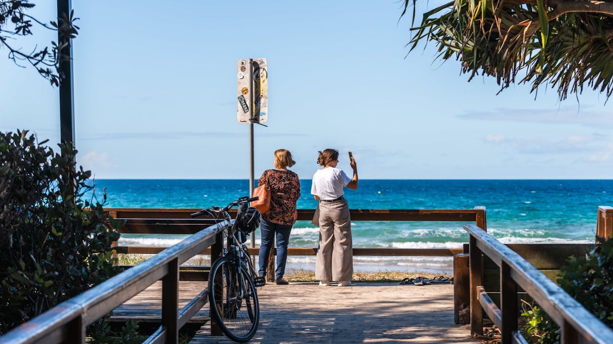 Shot of 2 women at the end of a small boardwalk with ocean in the background. Mudjimba Park entrance