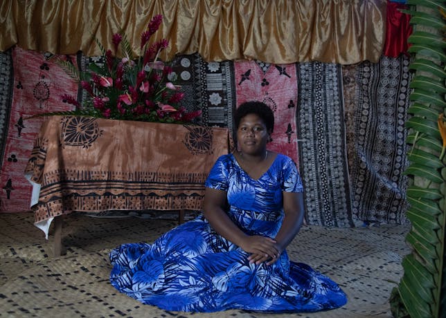 A photograph of a woman sitting down wearing a blue dress in front of multiple layers of fabrics