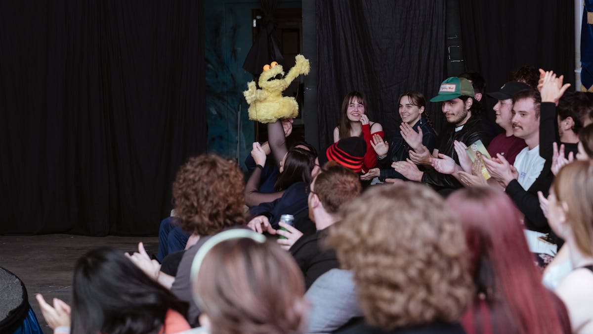 A crowd of people seated in rows applauds energetically. A puppet resembling a yellow bird is raised