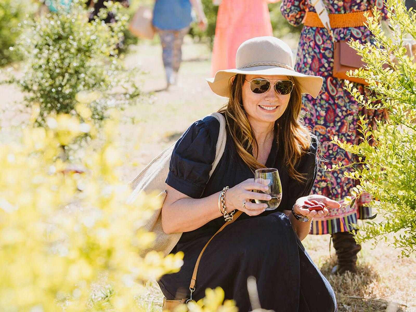 Lady crouching down in a sunny paddock smiling, holding a glass of wine and freshly picked fruit.