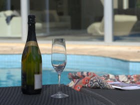 Bottle of sparkling wine next to champagne flute on pool lounge next to swimming pool
