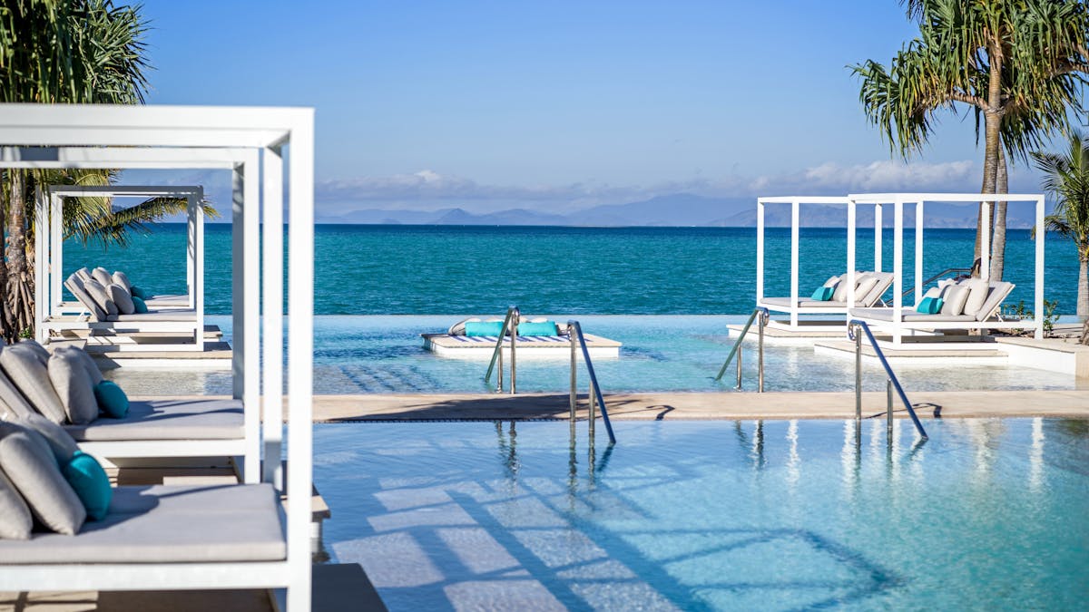 Infinity pool with white cabanas and turquoise cushions overlooking the ocean, framed by palm trees.