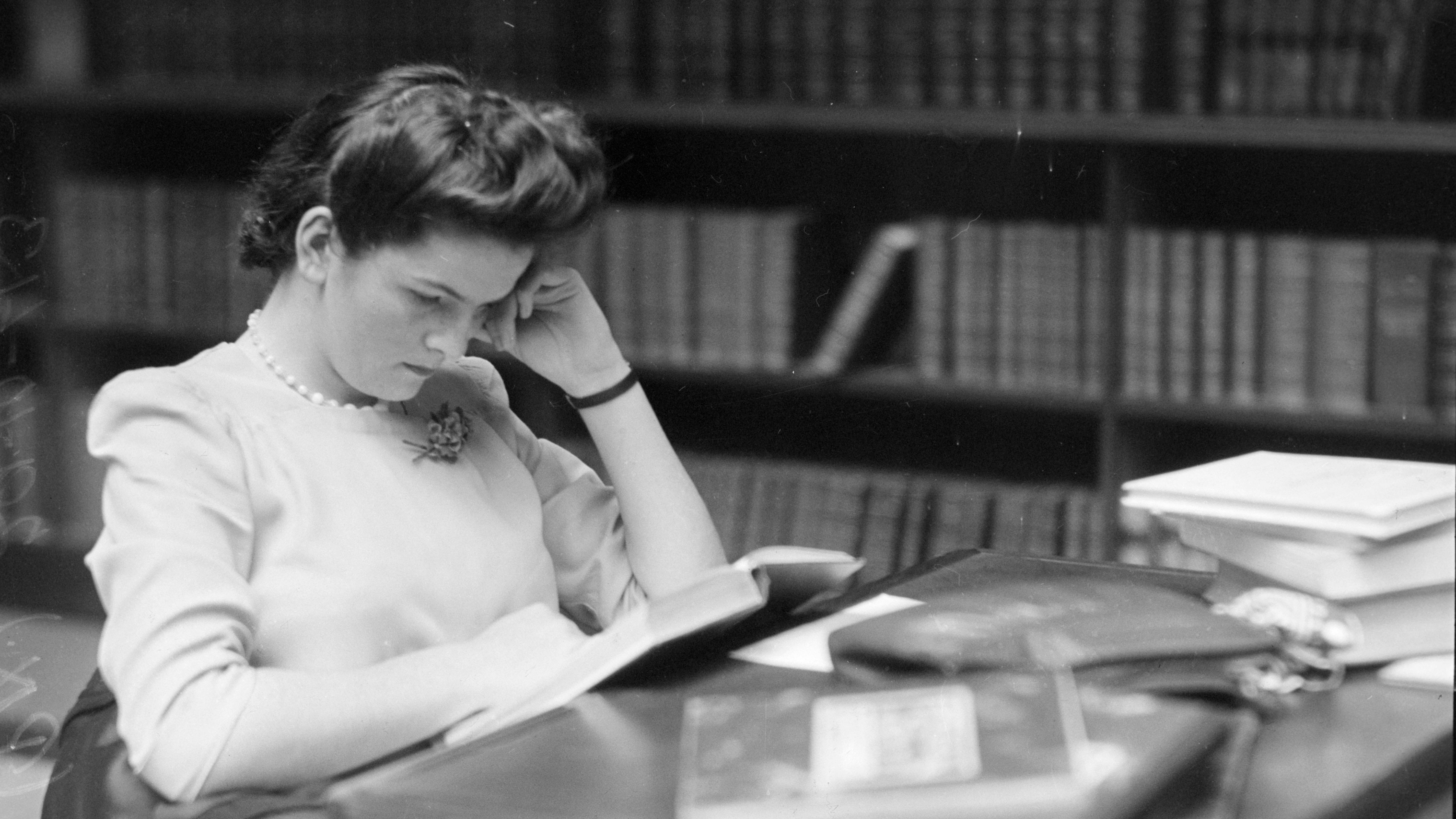 A black and white photo of a woman reading at a library desk
