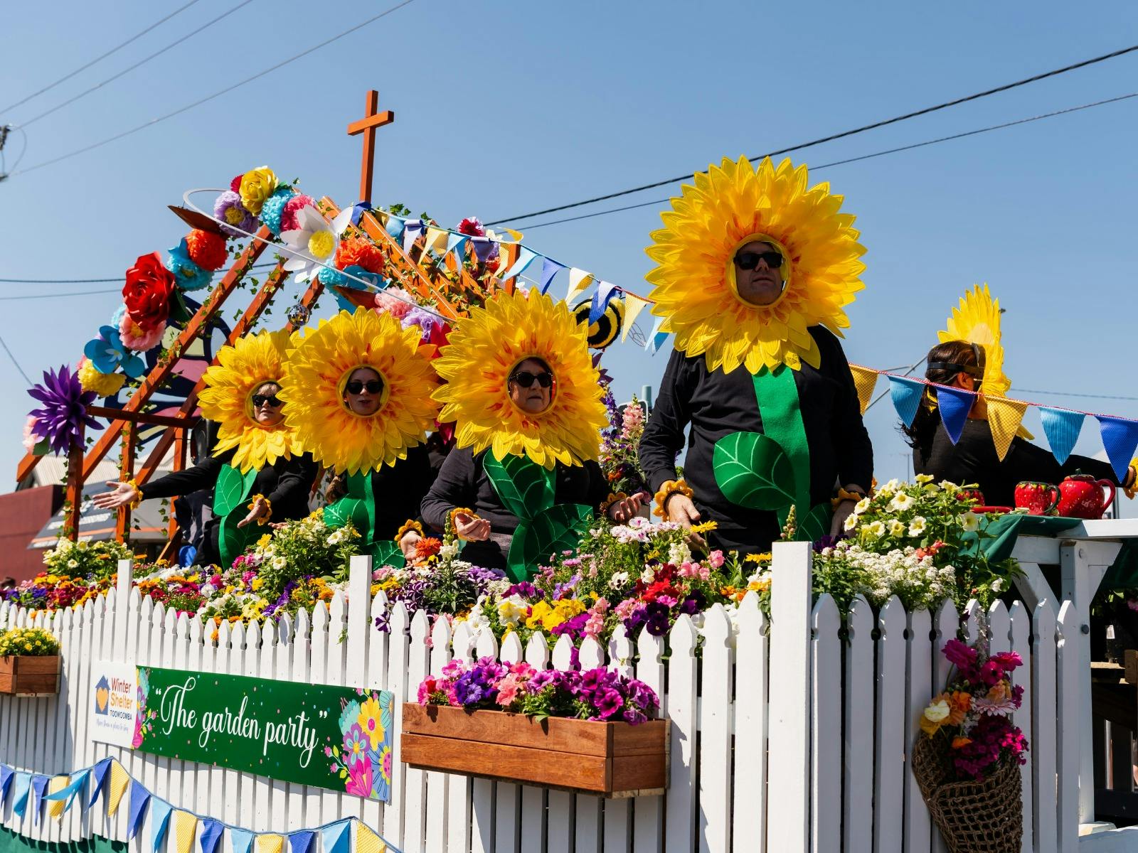 A group of men and women dressed as sunflowers sit on a parade float, behind white picket fencing.