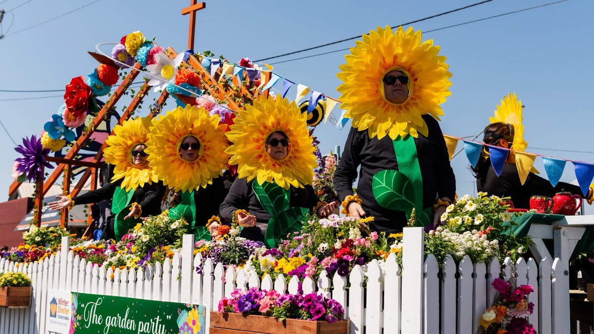 A group of men and women dressed as sunflowers sit on a parade float, behind white picket fencing.