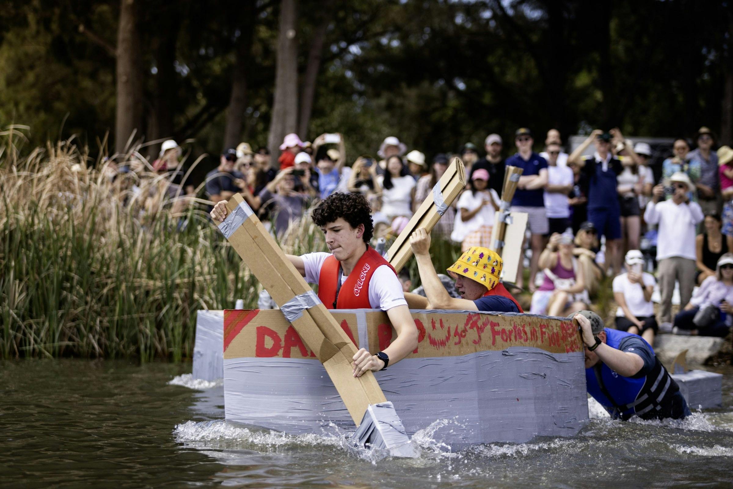 Young paddlers racing a cardboard boat on the lake while a crowd watches from the shore