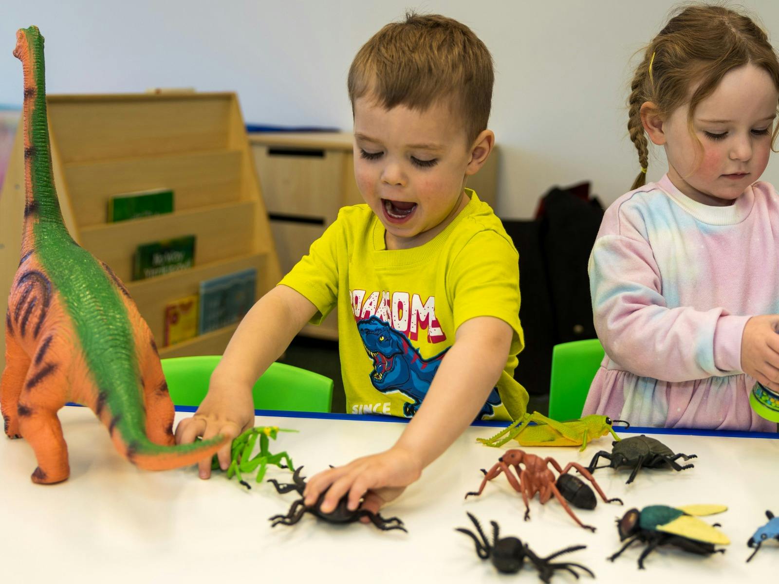 A young boy wearing a yellow shirt with blue dinosaur plays with toy dinosaurs at Science Time