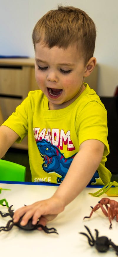 A young boy wearing a yellow shirt with blue dinosaur plays with toy dinosaurs at Science Time