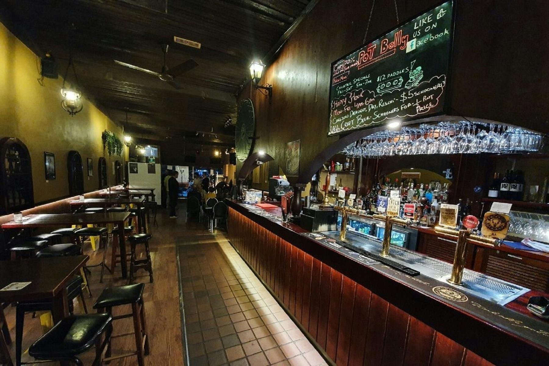 Row of bar tables and stools line the wall on the left, in front of a brightly lit bar on the right