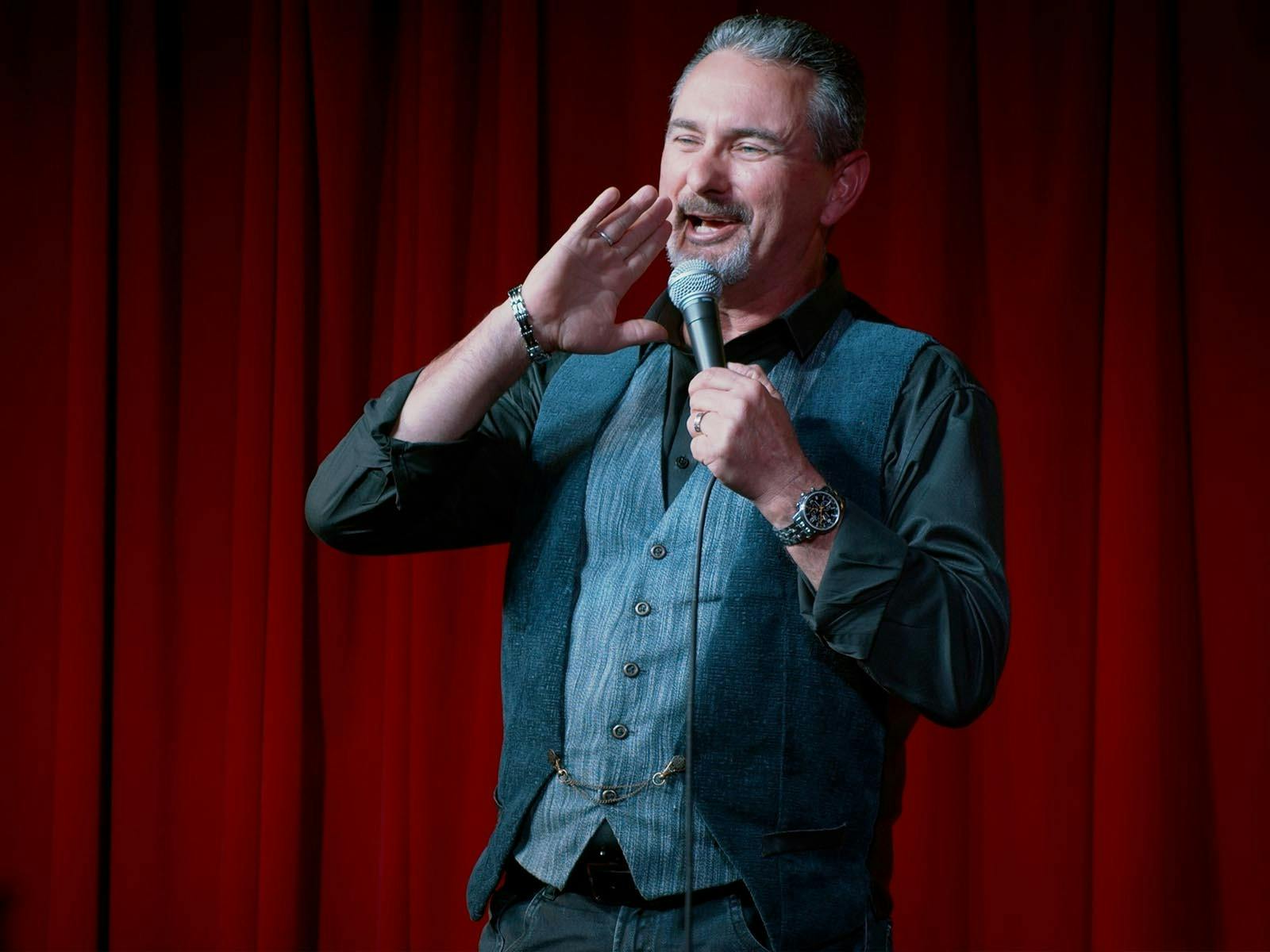 A white man in a grey waistcoat holding a mic