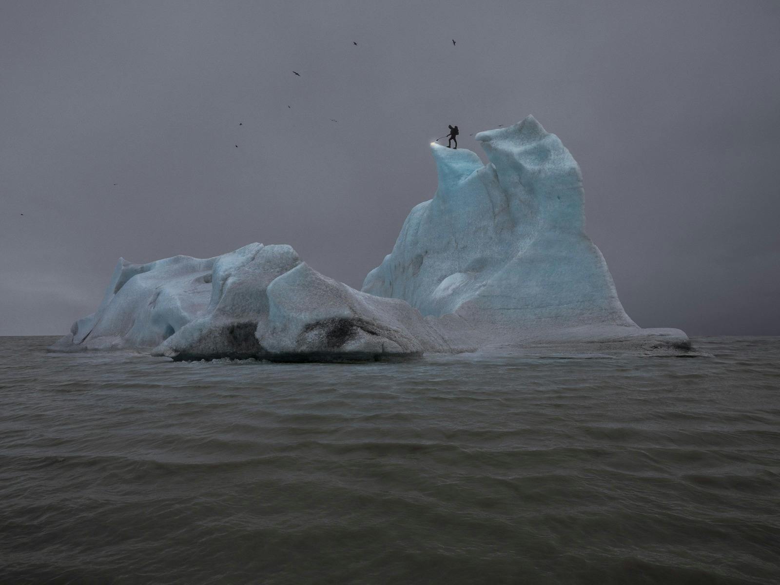 A person stands atop an iceberg, surrecounded by sea, with a blowtorch, setting fire to the ice.