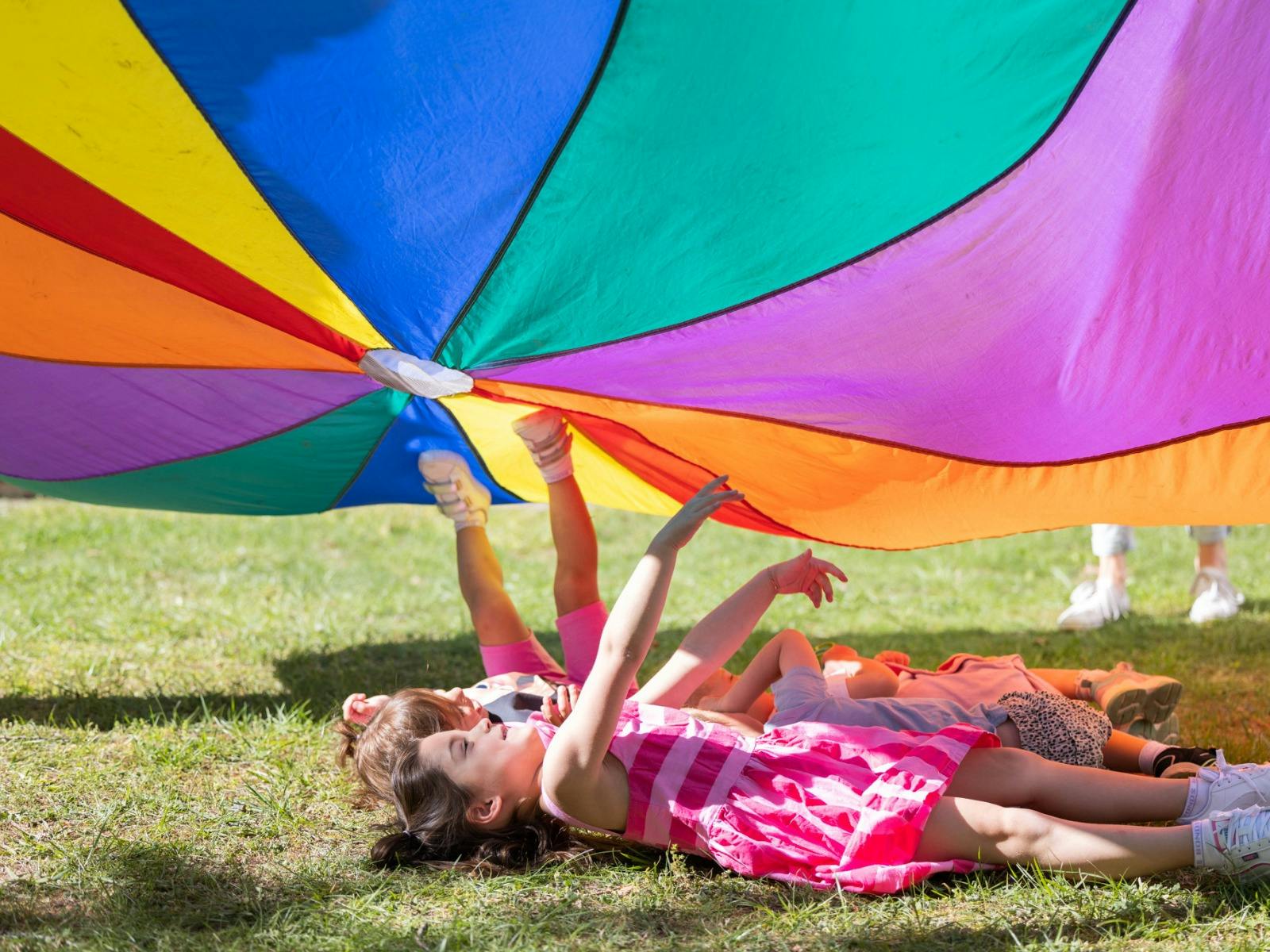 A group of children laying in the grass under a rainbow parachute.
