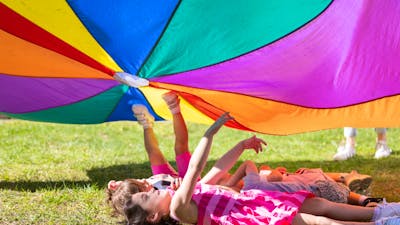A group of children laying in the grass under a rainbow parachute.