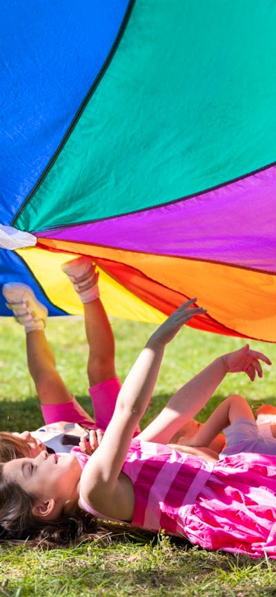 A group of children laying in the grass under a rainbow parachute.