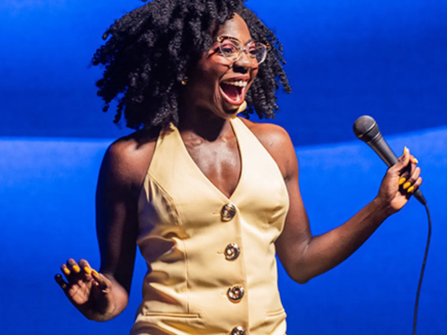 dark lady with long curly hair and glasses with microphone in right hand laughing