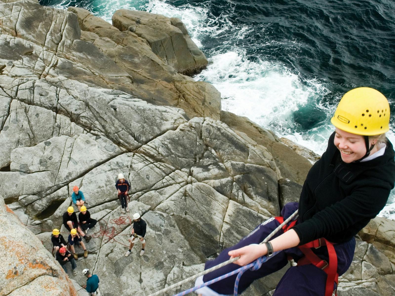 A woman abseils down a rocky cliff by the sea. A group of abseilers sit on the rocks below.