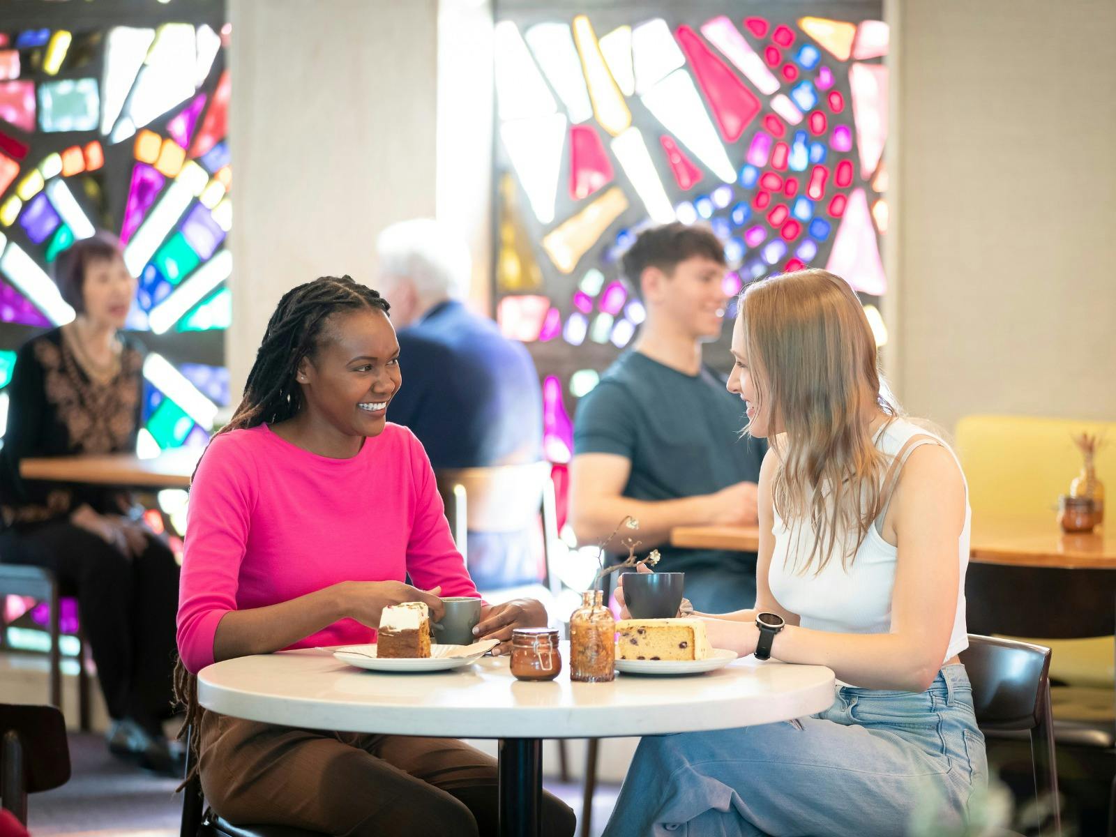 Two young women in a brightly lit cafe, sitting at a white table.