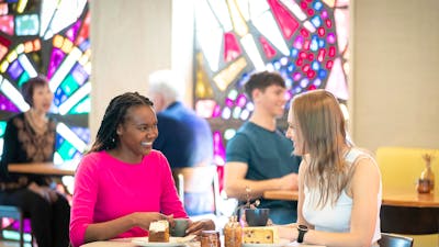Two young women in a brightly lit cafe, sitting at a white table.