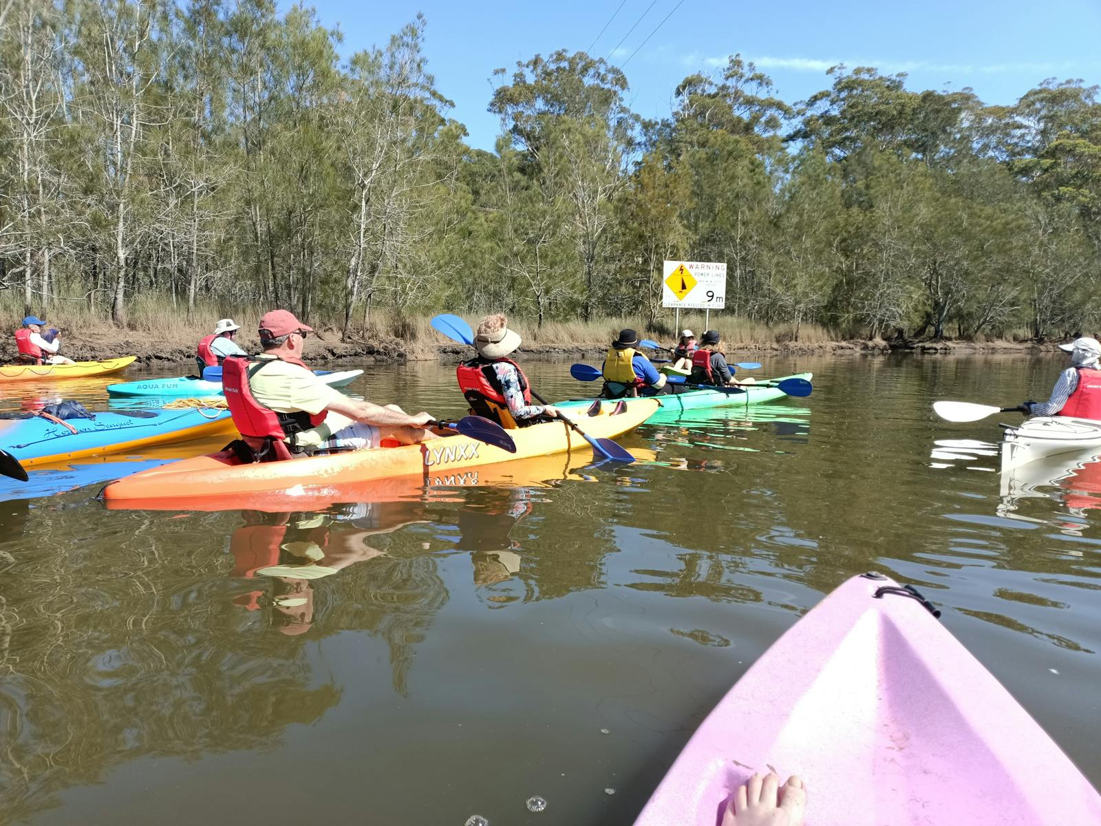 People paddling colourful kayaks witha clear sky and trees along the bank
