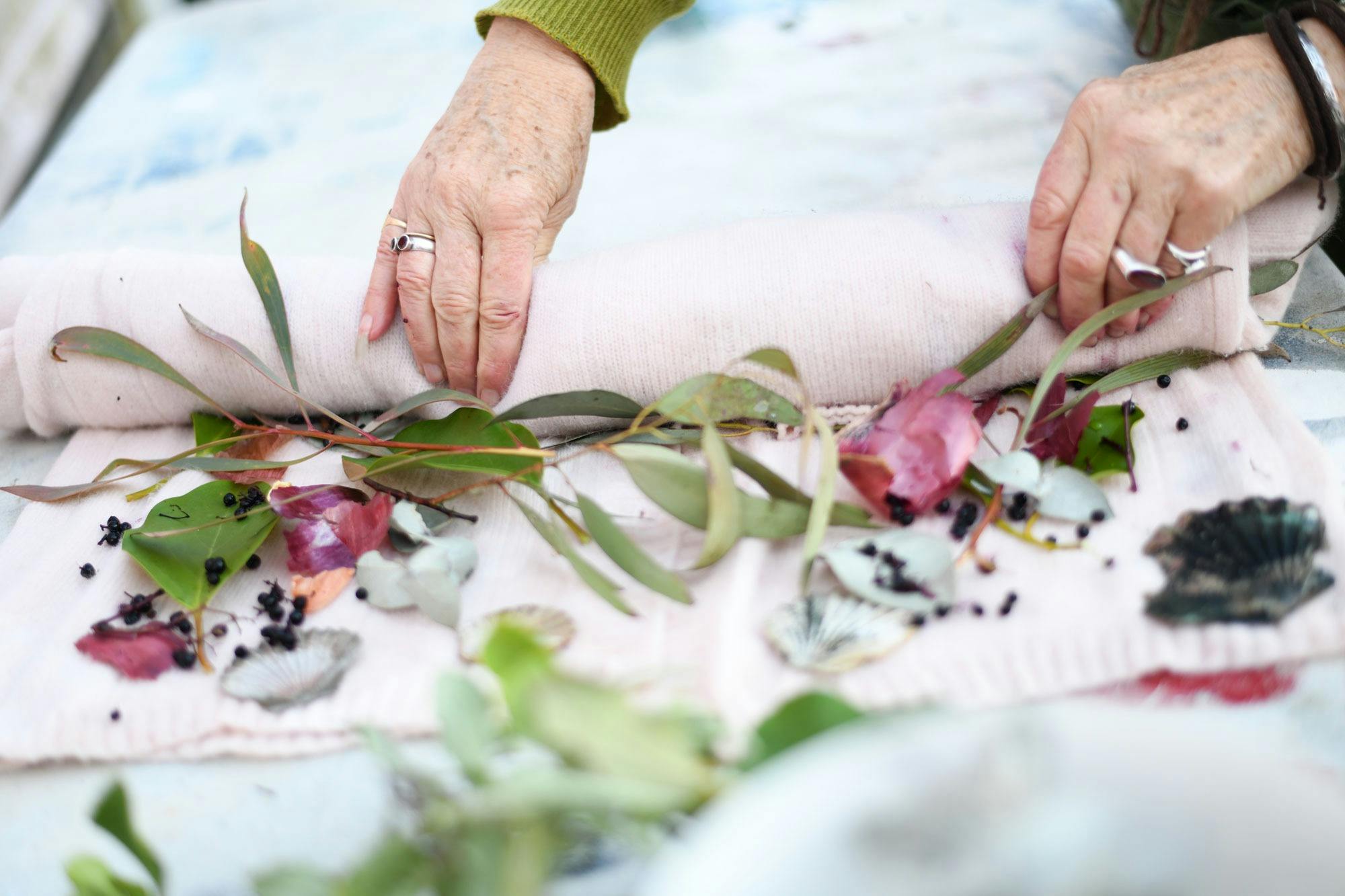 Close-up of hands arranging leaves and flowers on pale pink fabric for natural dyeing.