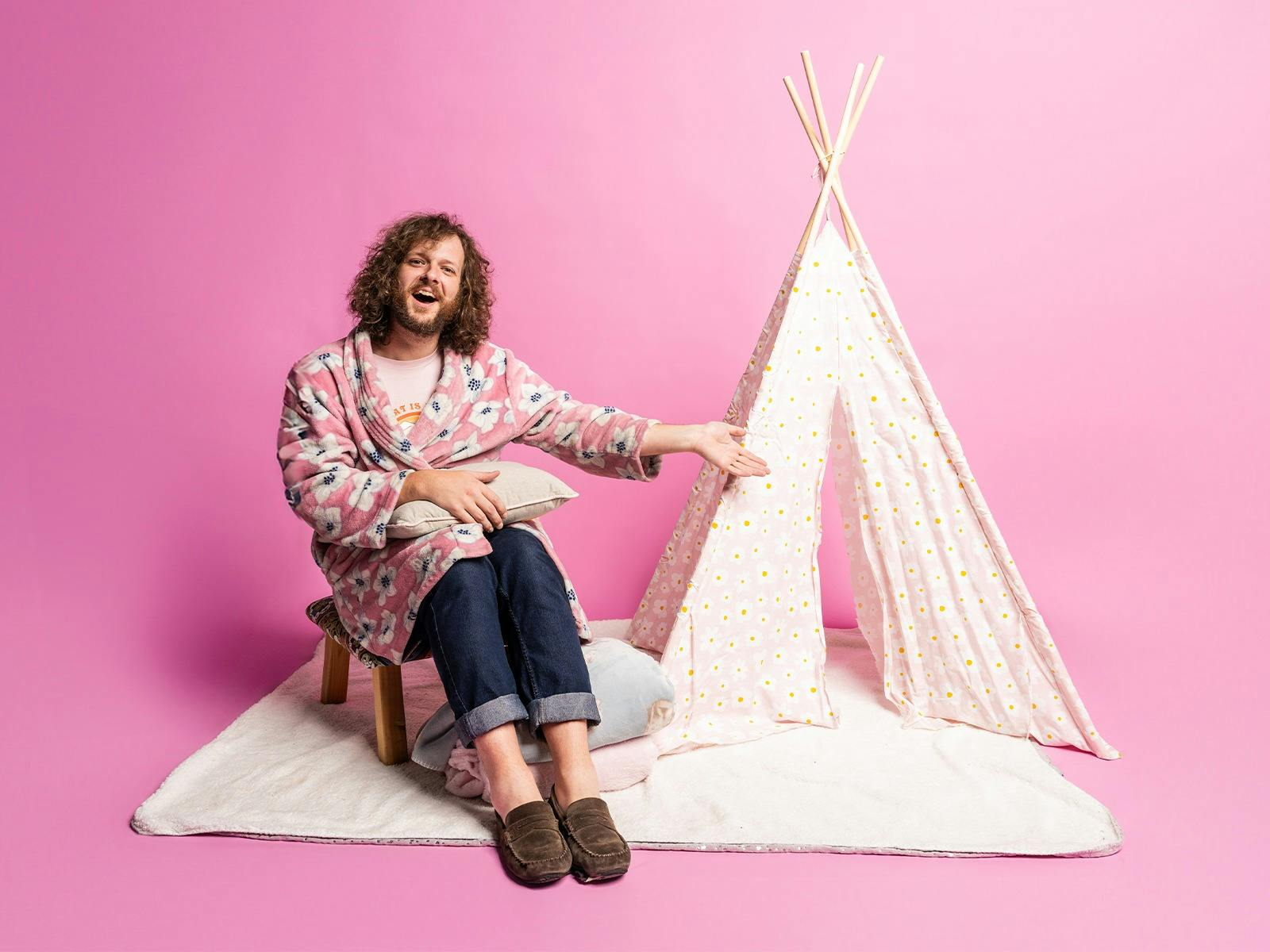 Blake sits on a stool by a child's tent, extending out his arm to welcome you in