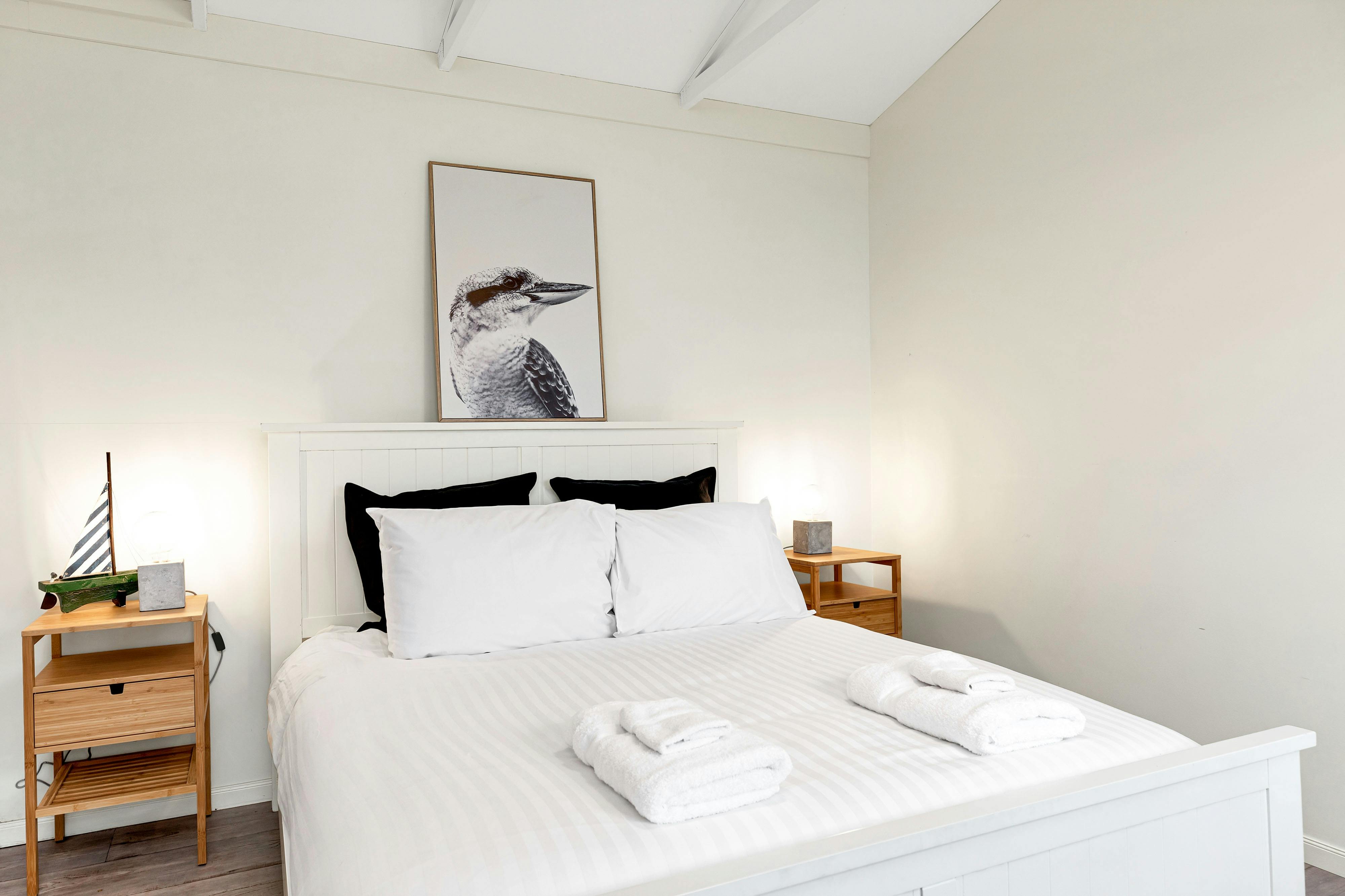 Queen sized bed made up with white linen, with framed picture of kookaburra, teak bedside tables