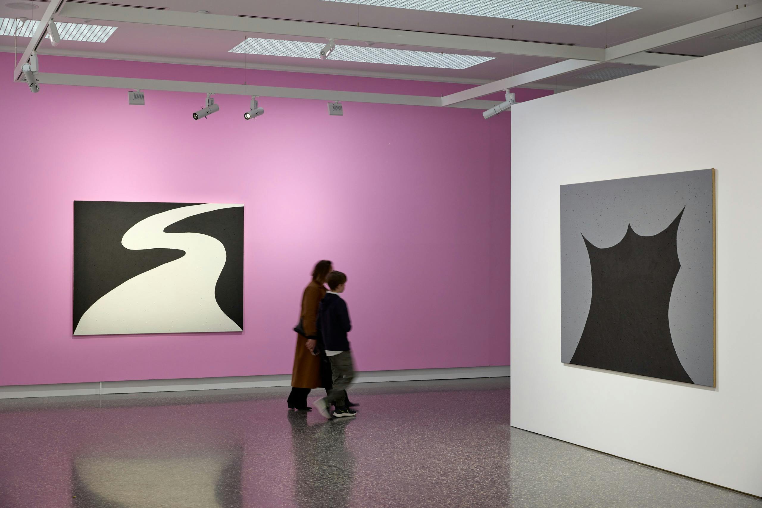 A gallery space with a pink wall, two people walk through the space
