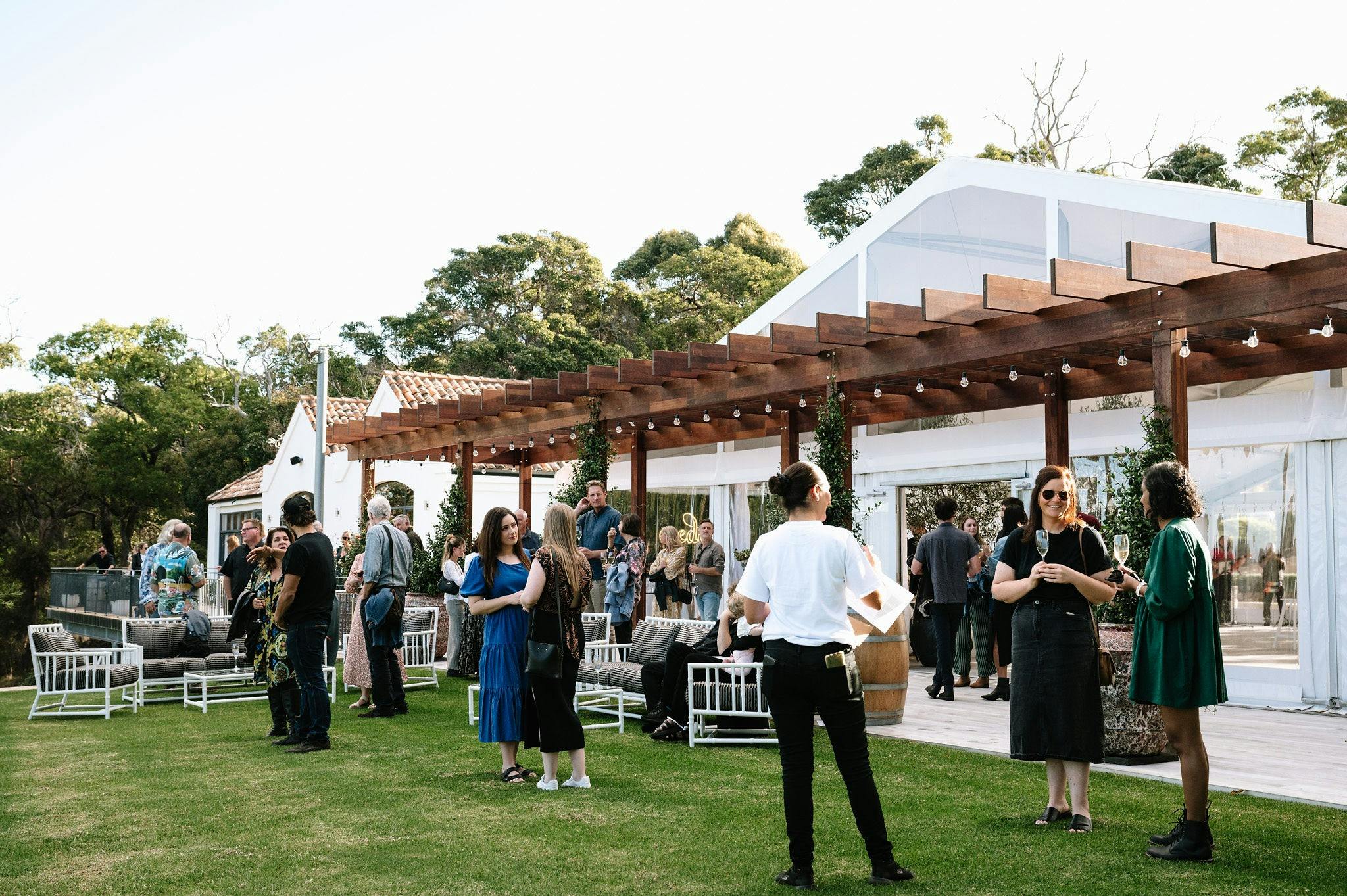 People gathering on grassed area outside large white marquee with wooden pergola