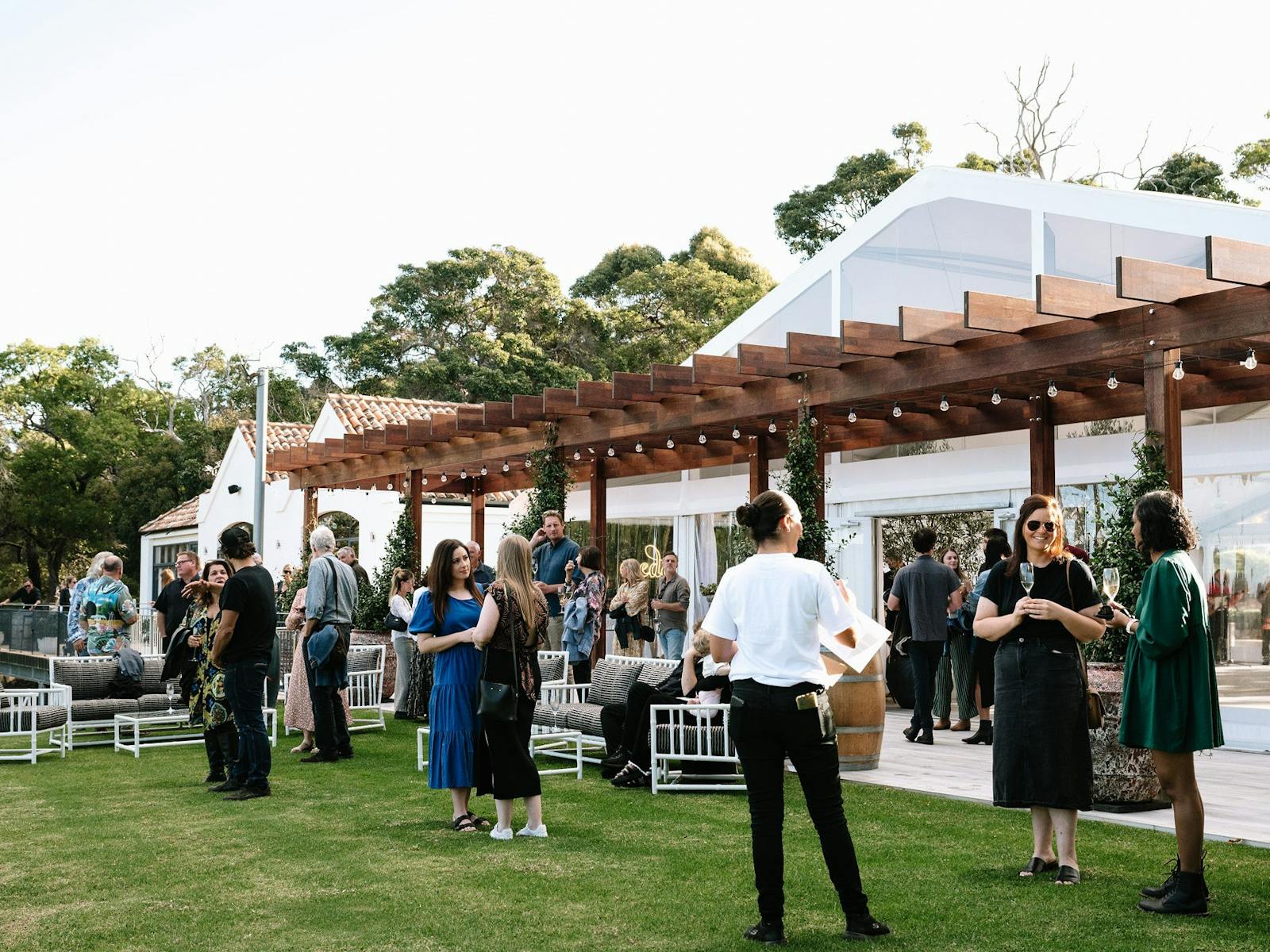 People gathering on grassed area outside large white marquee with wooden pergola