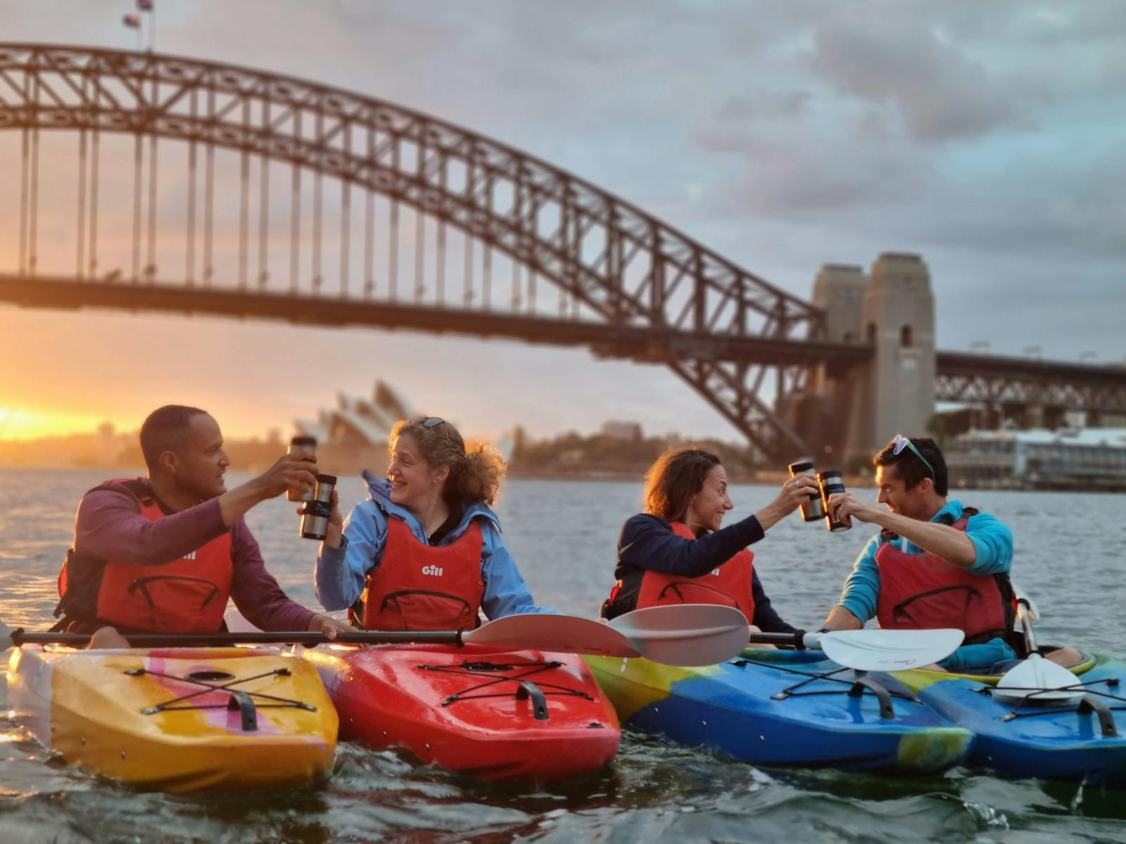four kaykers cheersing their coffees from kayaks in fron of sydney opera house and the harbor bridge