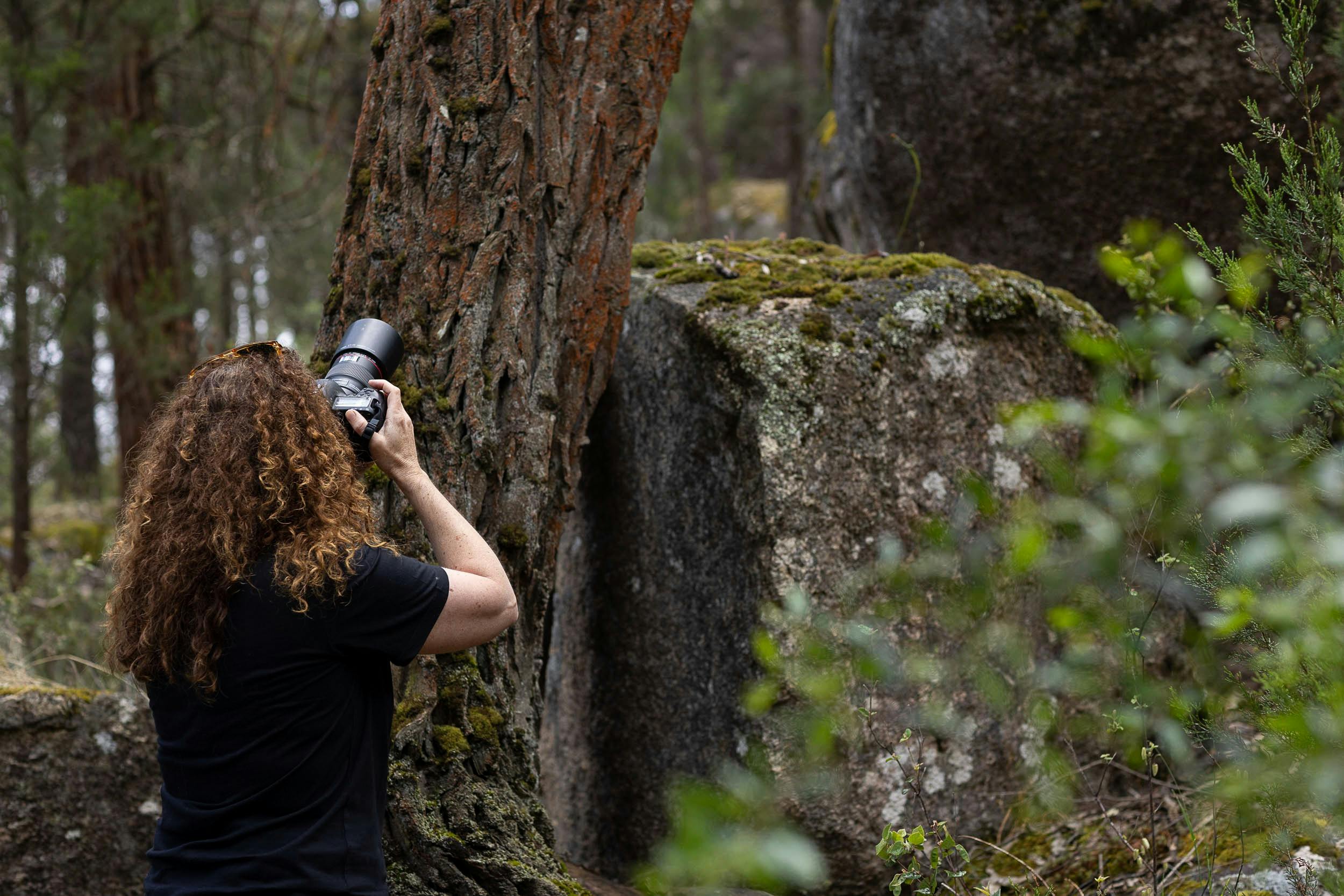 a woman with curly hair stands with back to camera holding a camera taking photos in the forest