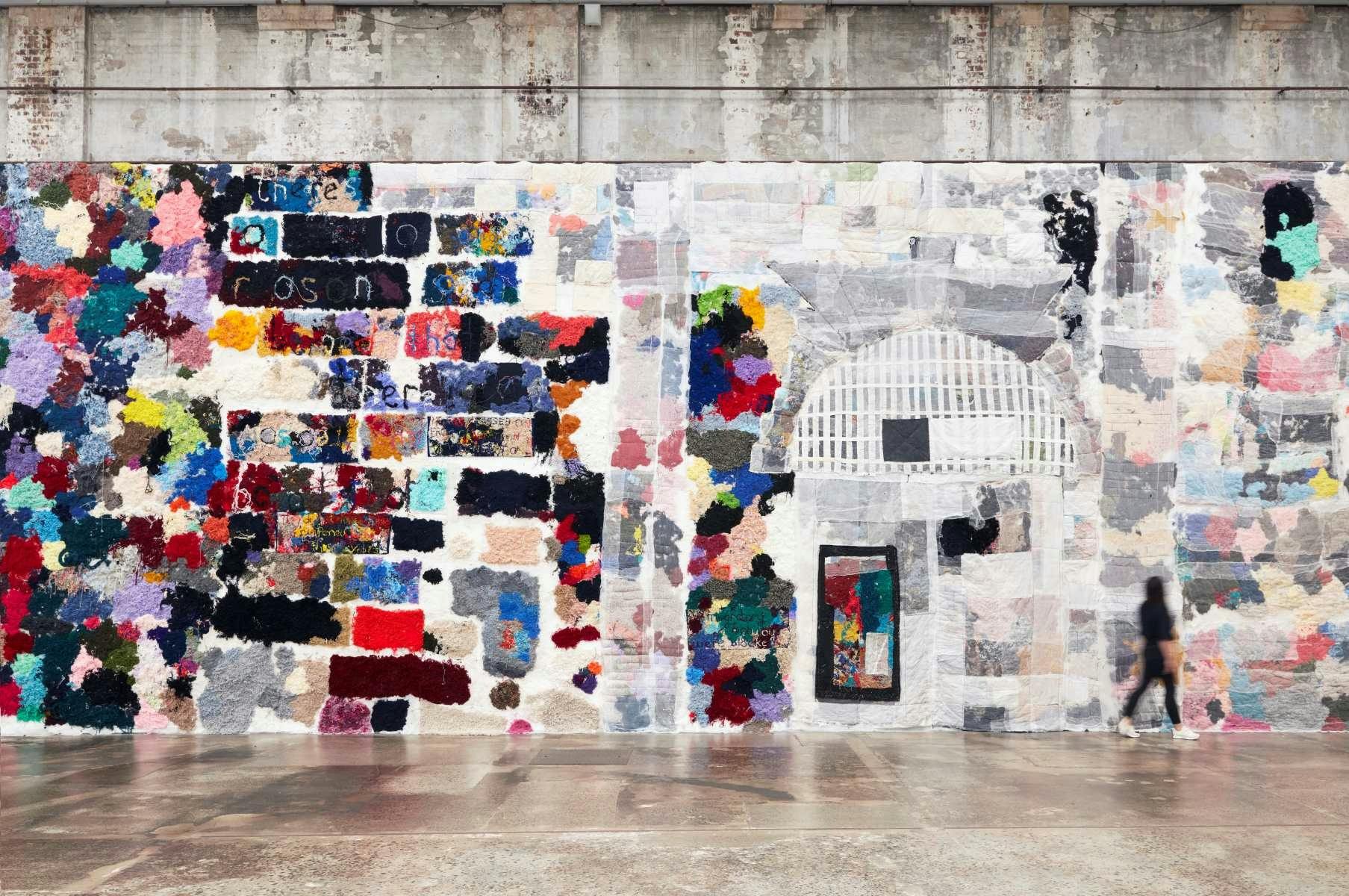 an exposed concrete wall covered in colourful patches of wool in the shape of big bricks.