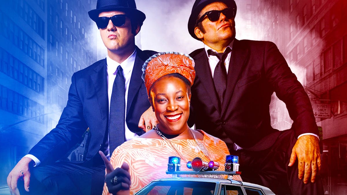 Two men dressed in matching suits, sunglasses and hats stand behind a woman in an orange dress