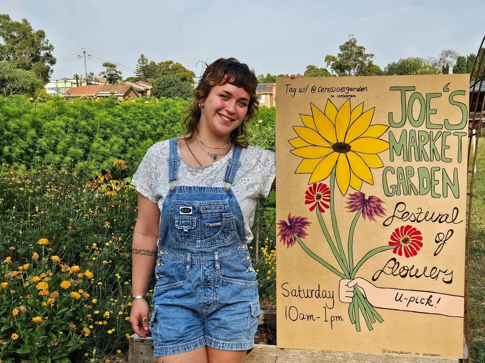 A person stands next to handmade sign which says Festival of Flowers on it