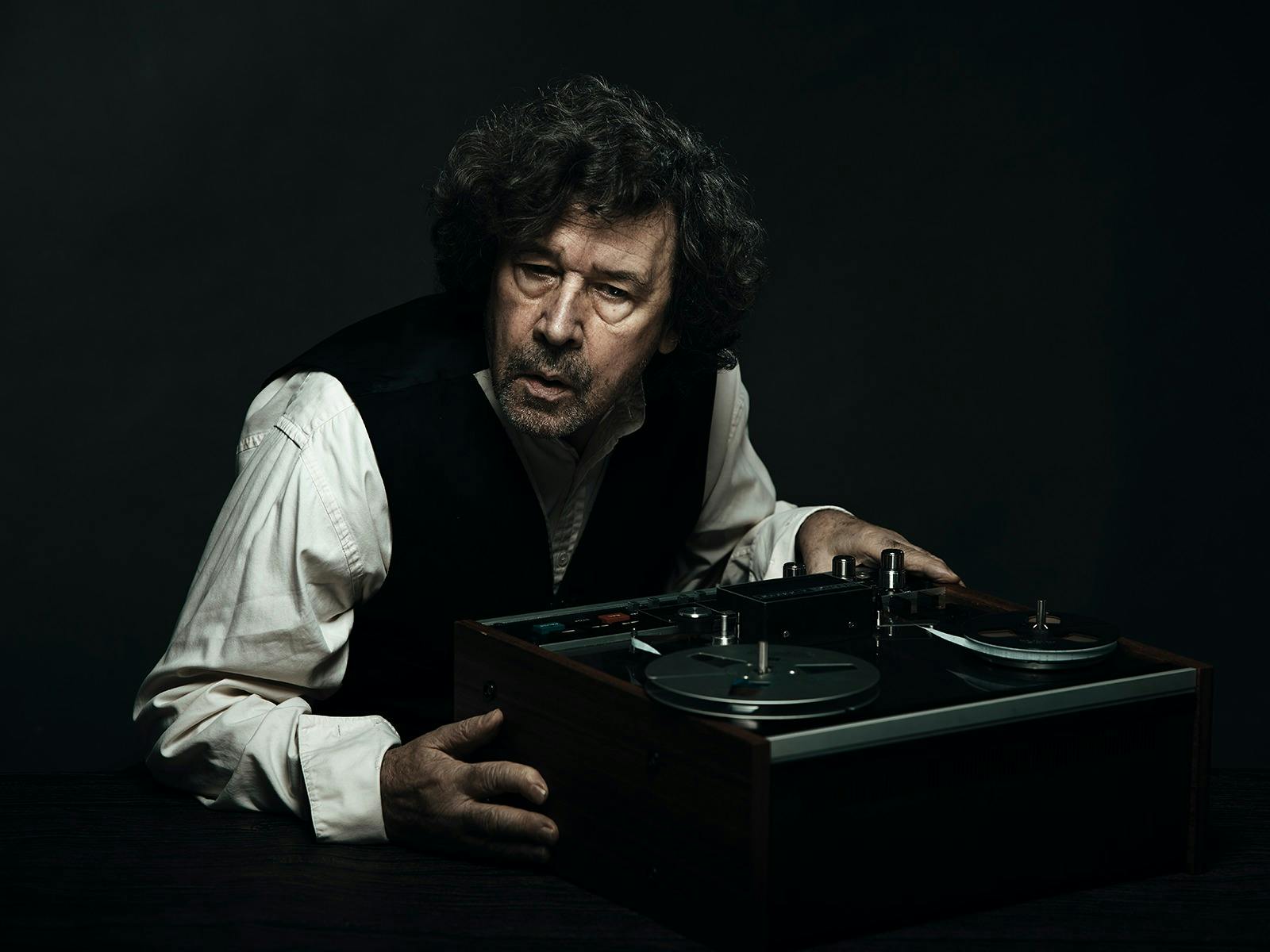 A man in a black vest and a white shirt in front of a black background leans over a tape recorder.