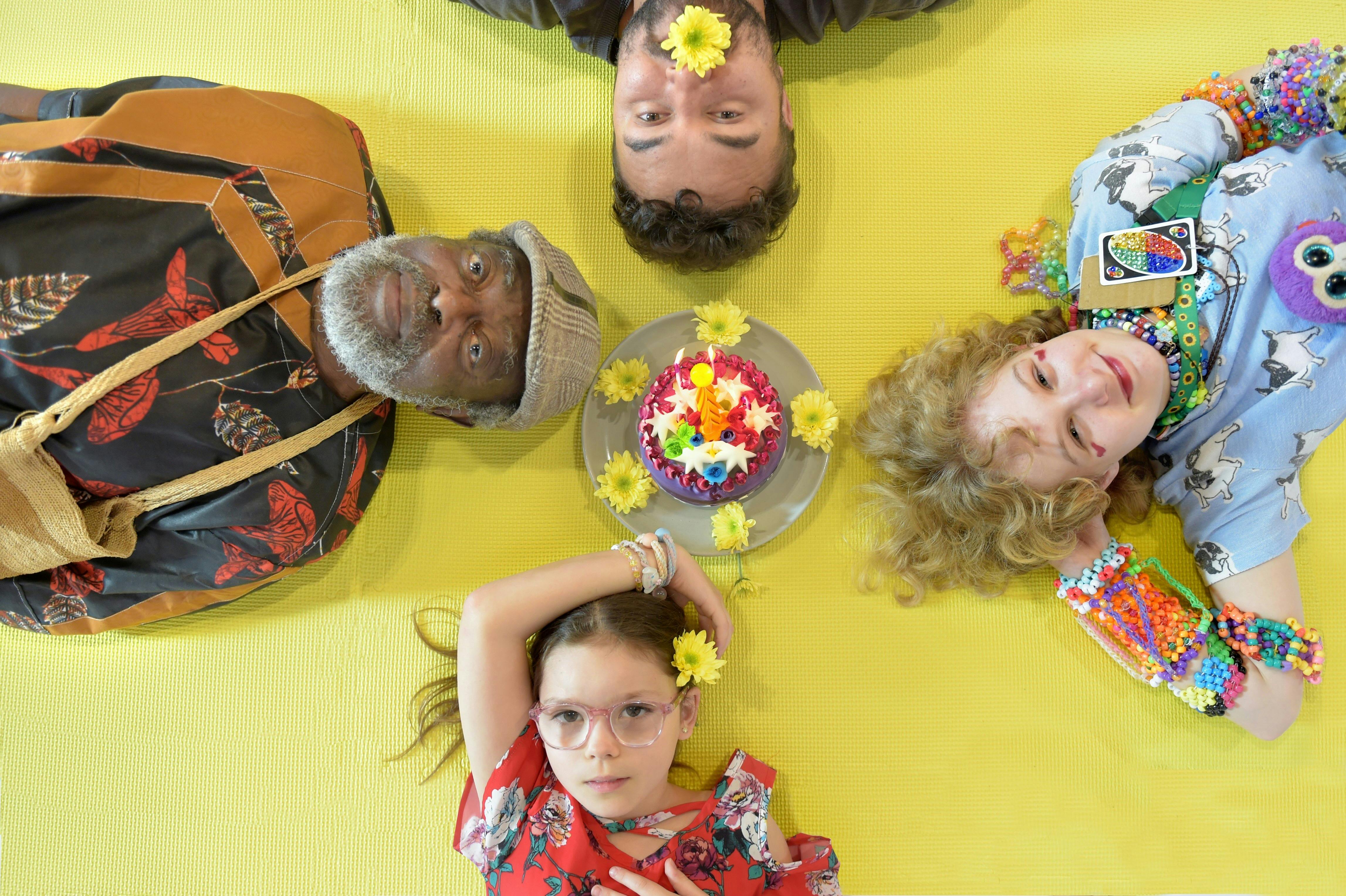 Four people across different ages look up at the camera while laying on a yellow background