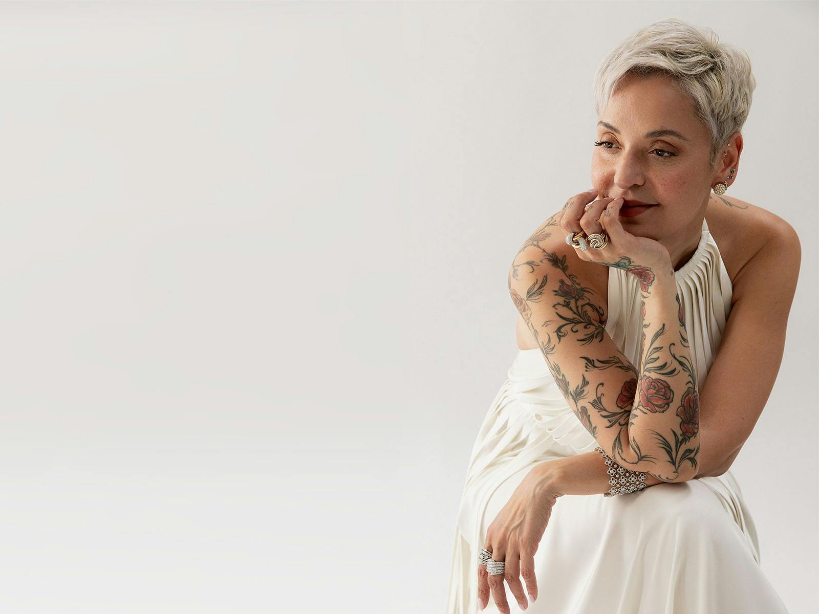 Mariza crouching in a white long dress in front of a white backdrop.. Her chin leaning on her hand.