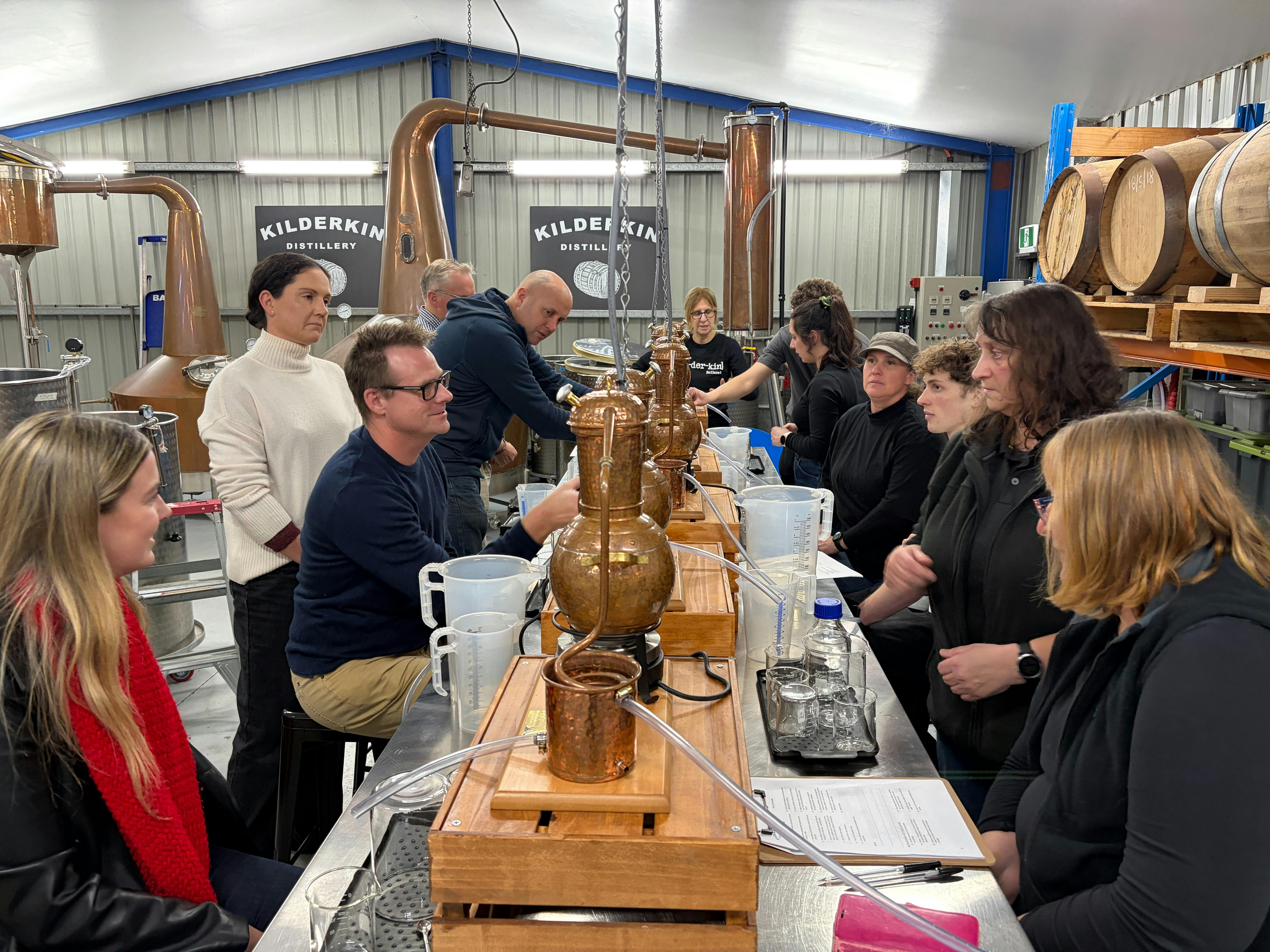 Ten adults sitting at bench with small stills distilling gin